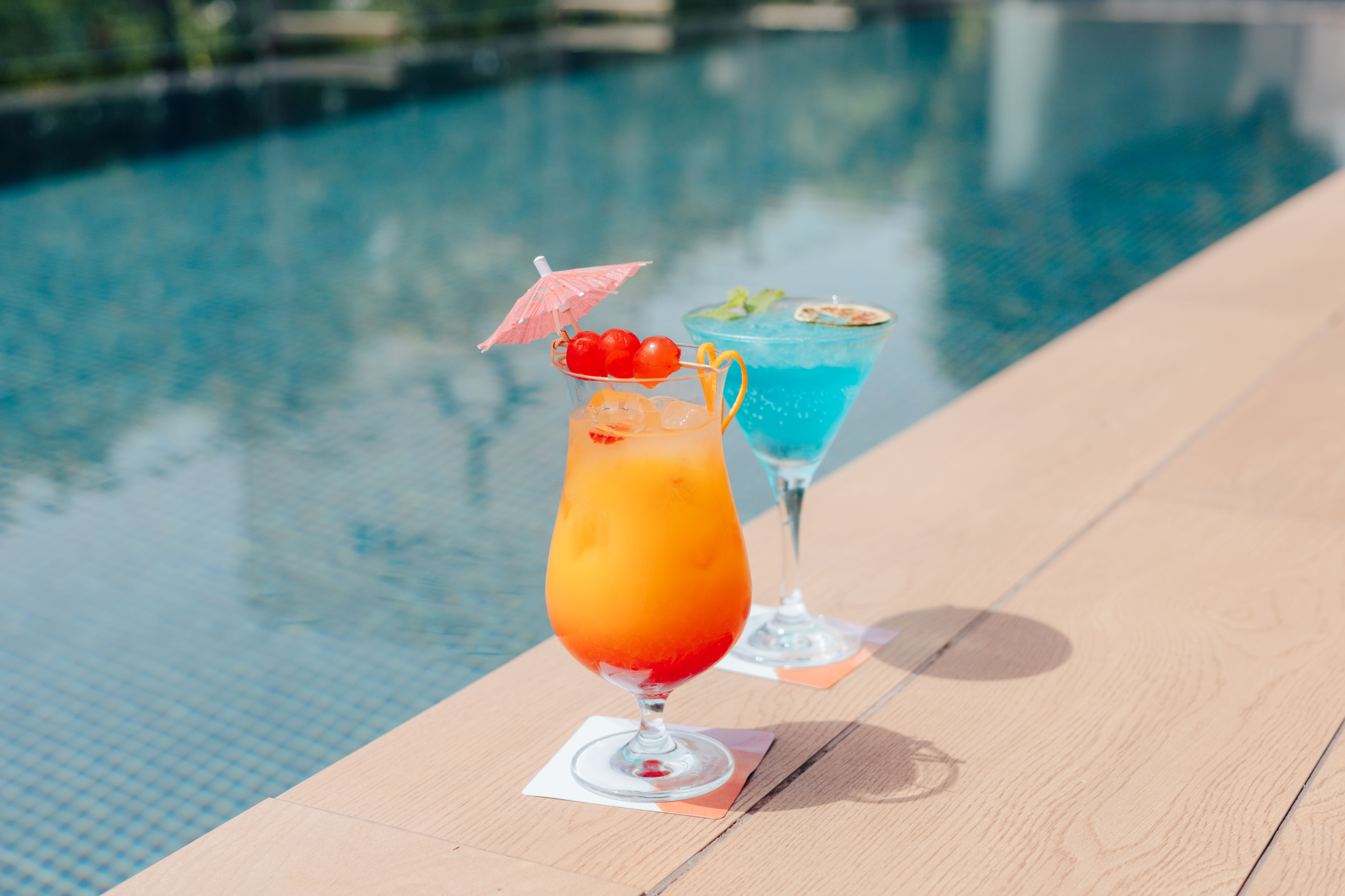Orange and blue tropical cocktails with garnishes by a swimming pool.