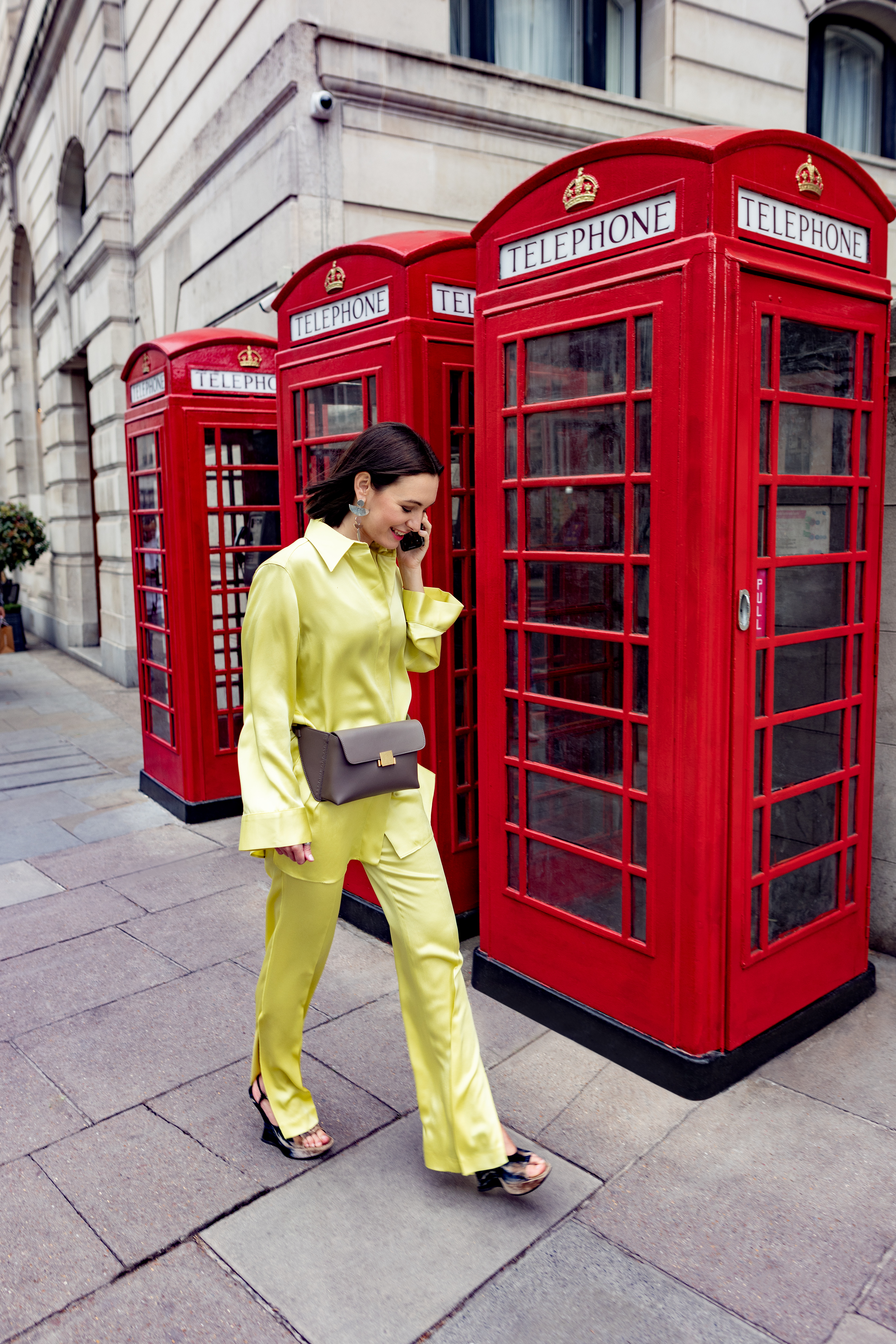A woman in yellow pants and high heels walks past red telephone booths.