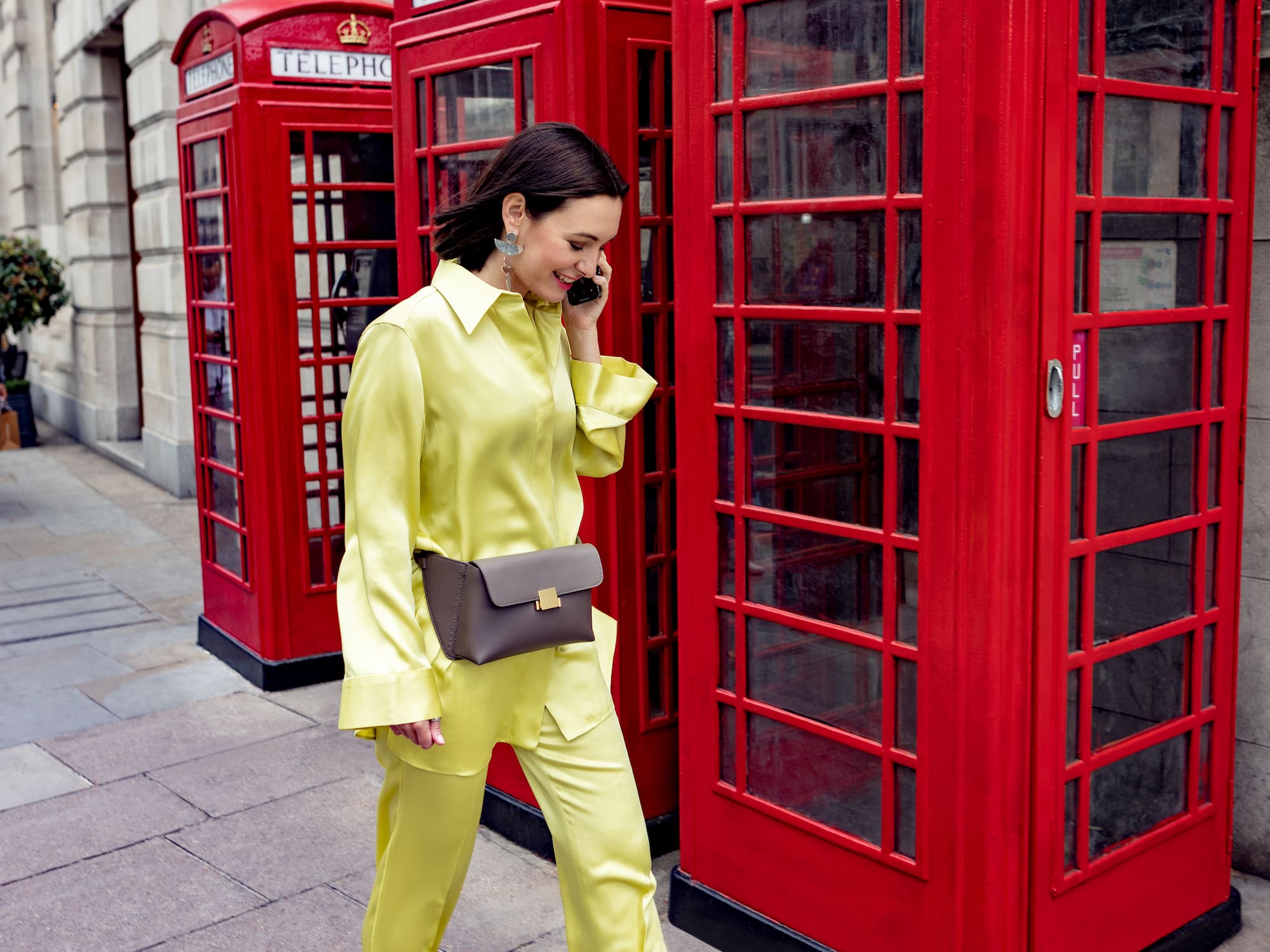 A woman in yellow pants and high heels walks past red telephone booths.