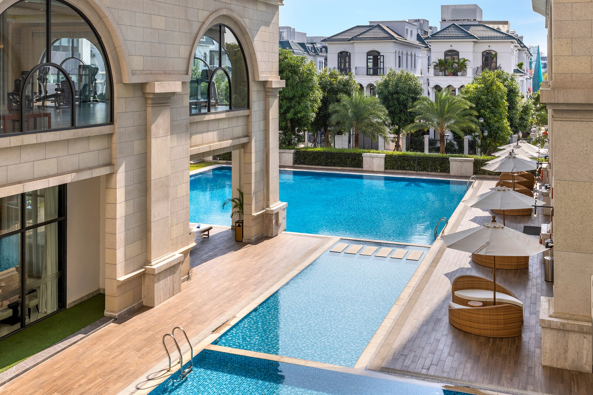 Luxury outdoor pool with gym view, modern villas, and shaded lounge chairs.