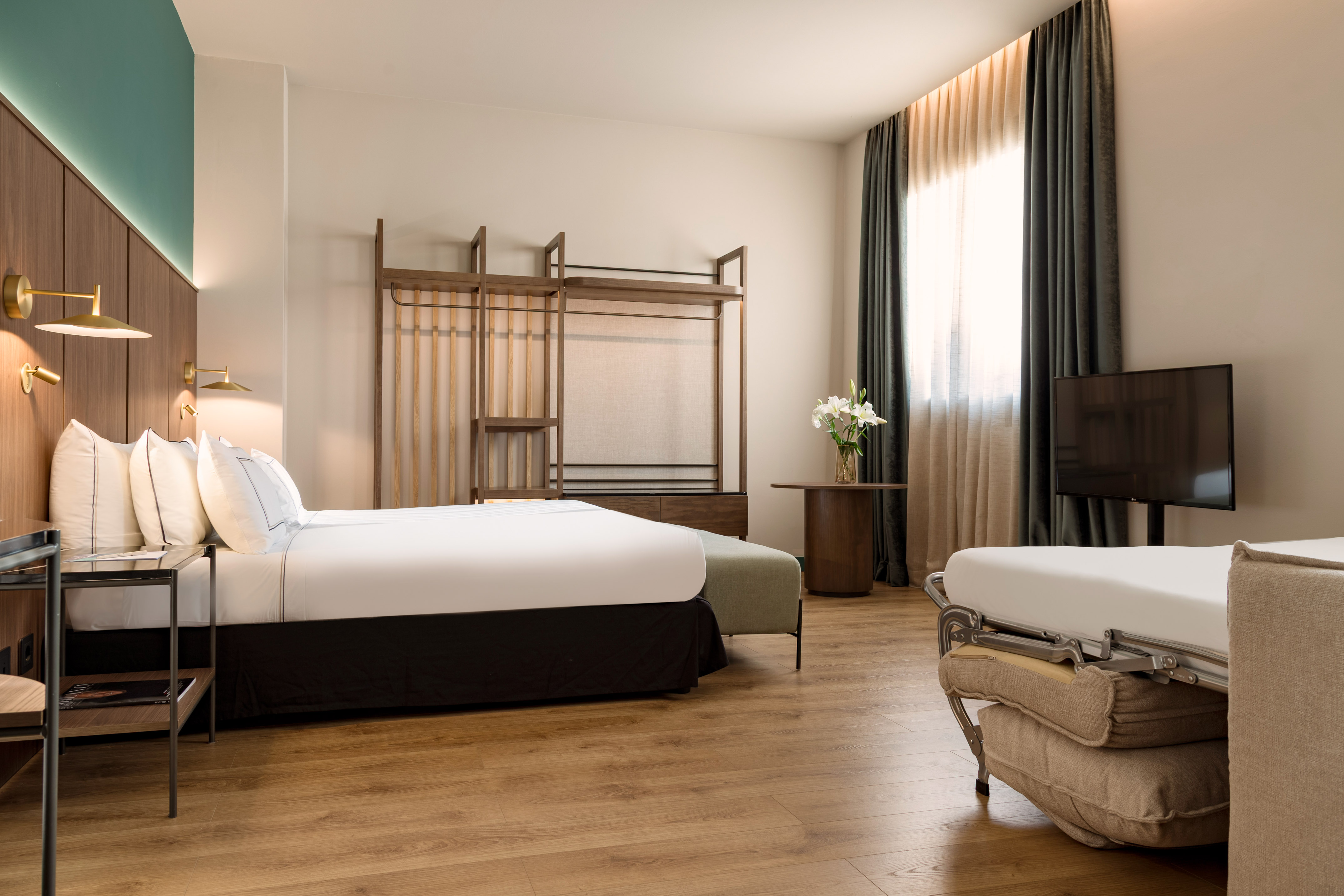 Modern hotel bedroom with king bed, wood floor, teal accent wall, open closet, and TV.