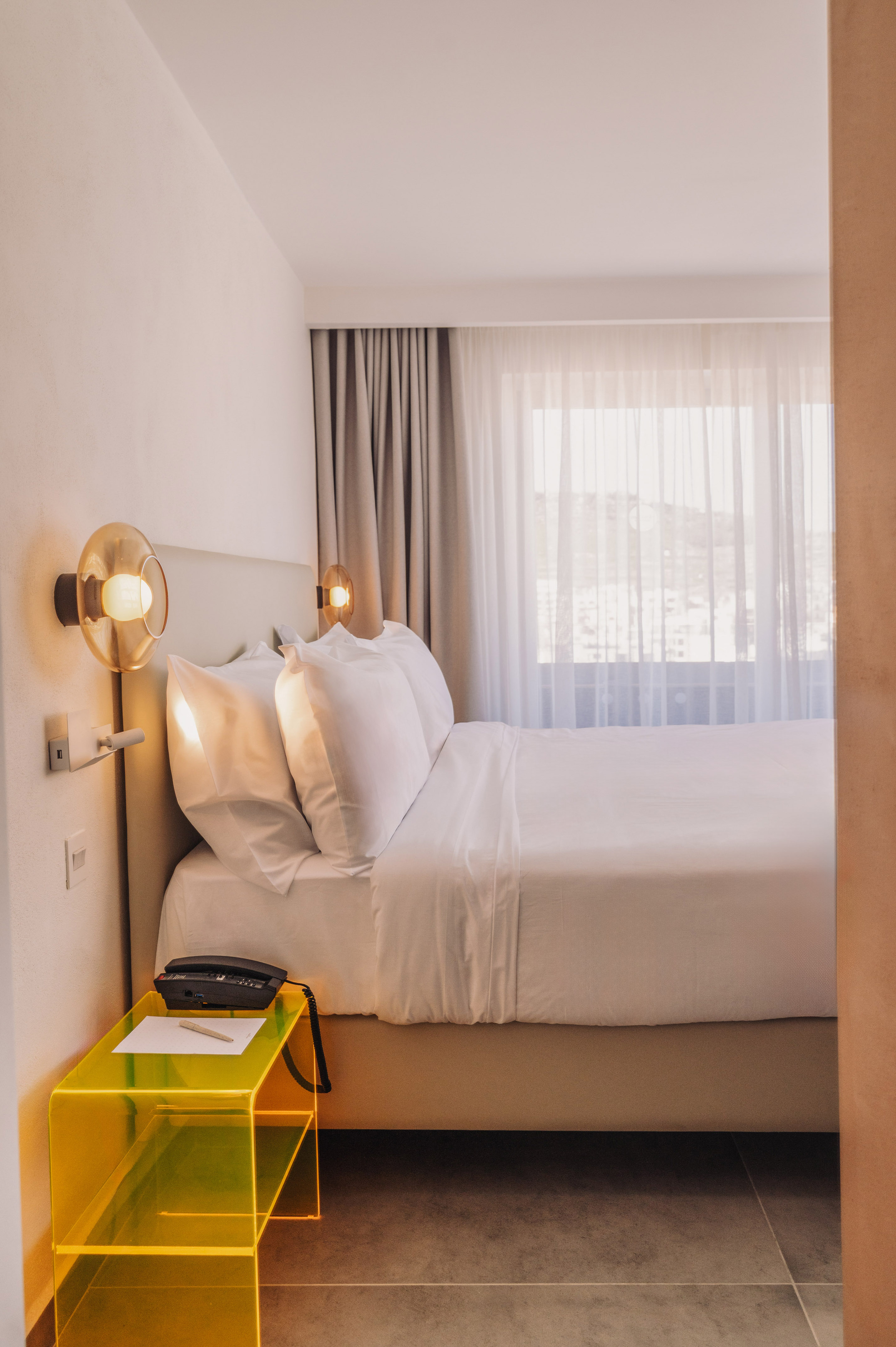 Modern hotel bedroom with white bed, warm bedside lamps, yellow acrylic nightstand.