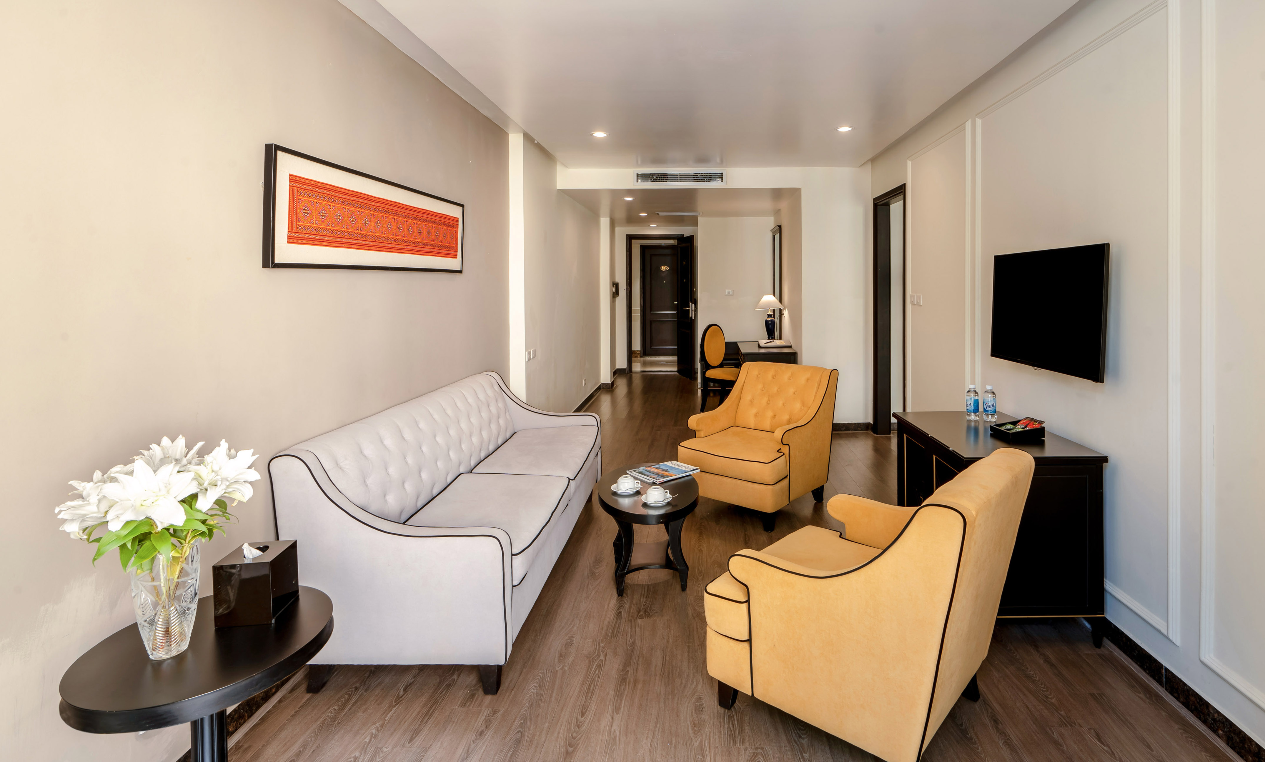 Hotel suite living area with light gray sofa, yellow armchairs, and TV.