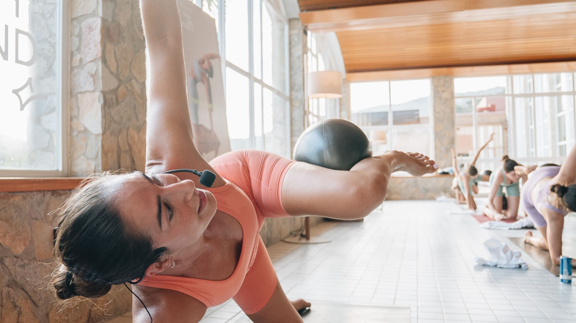 Woman exercising side plank with stability ball in a sunny yoga studio.