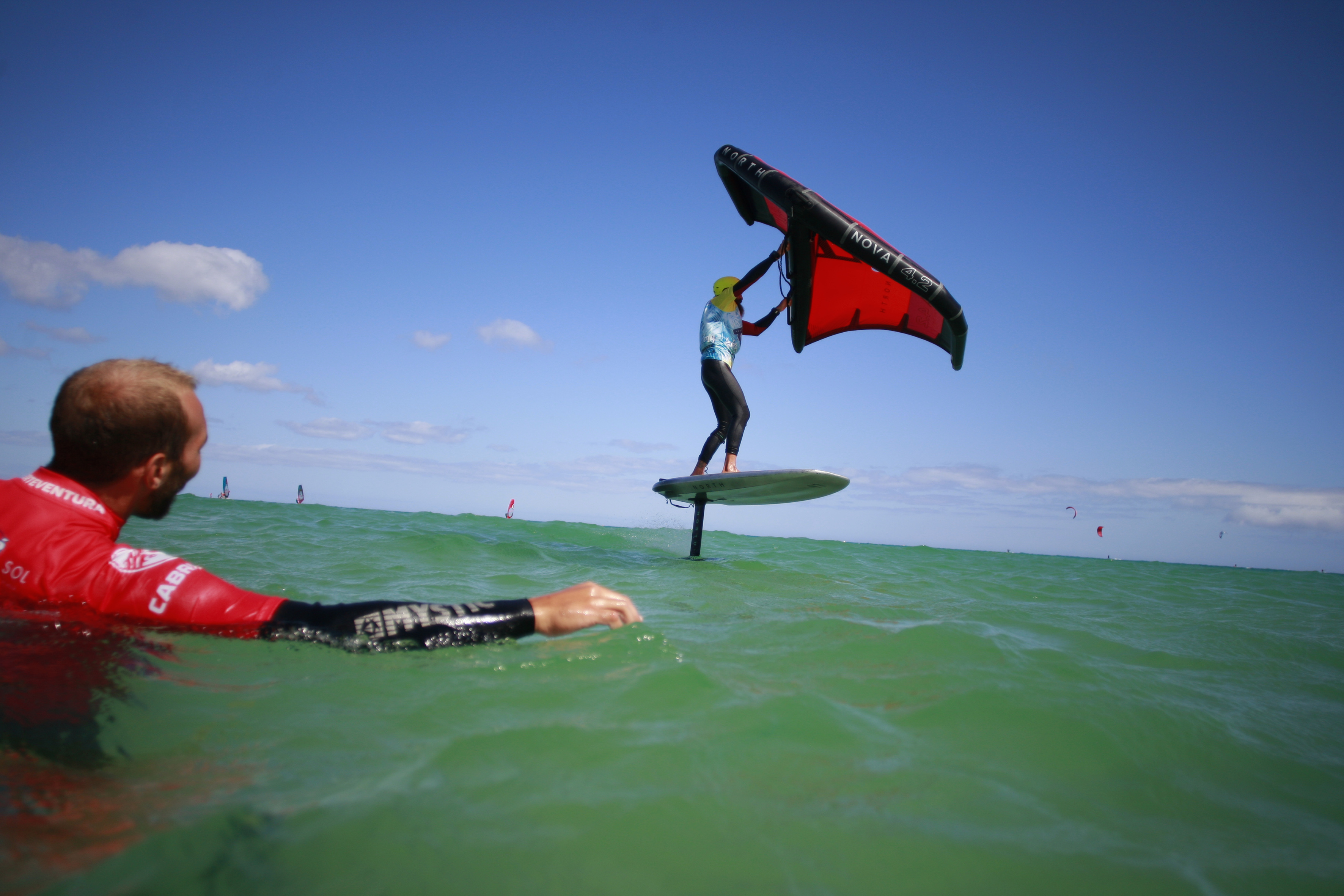 Wing foiler on hydrofoil board with red wing sail, watched by man in green water.
