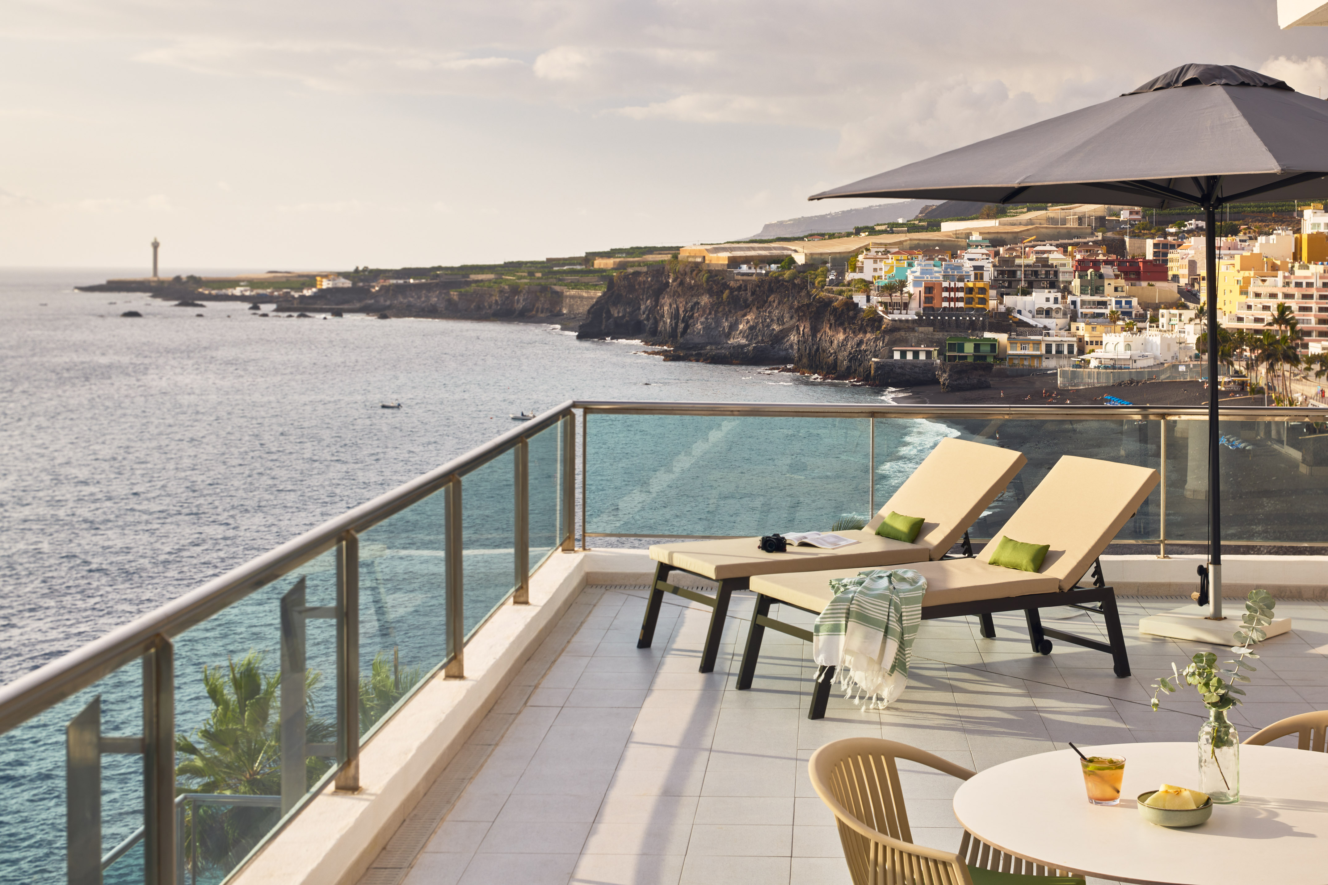 Resort balcony with loungers, ocean view, colorful coastal town, and lighthouse.