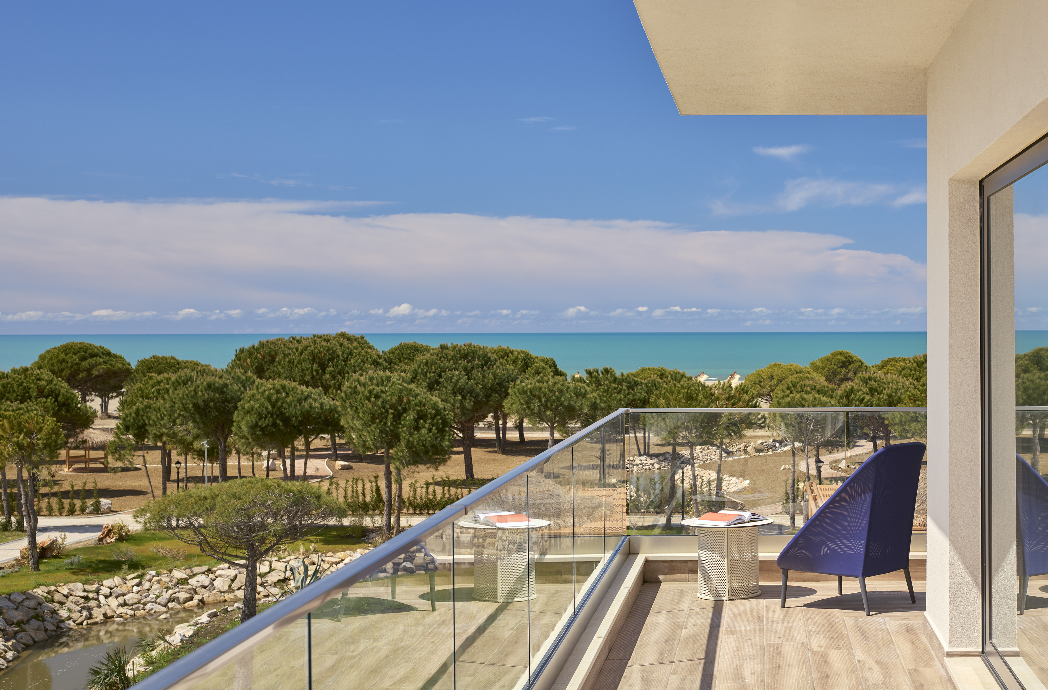 Hotel balcony with sea view, lush pine trees, and sunny blue sky.