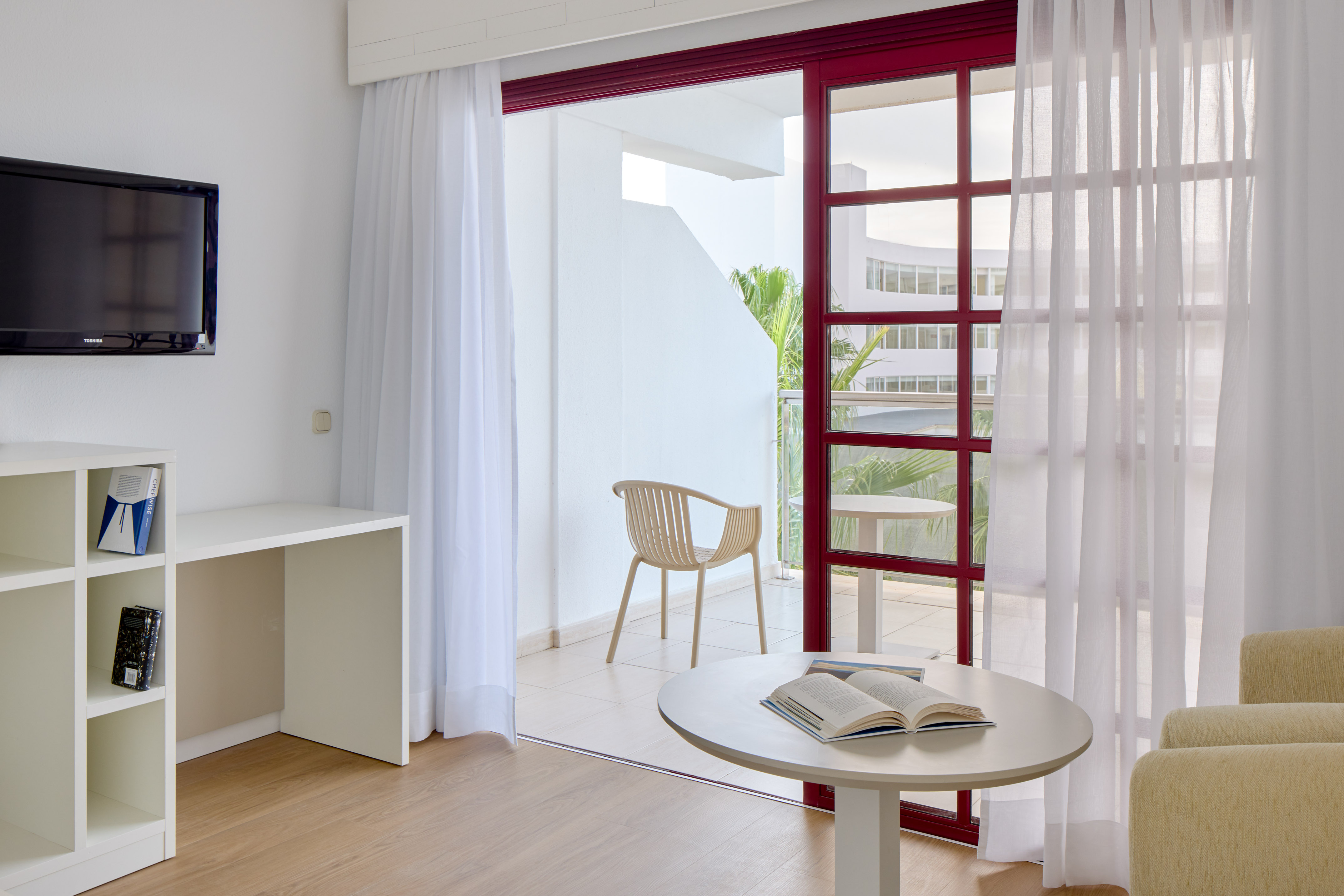 Hotel room with balcony, TV, white desk, and light wood floor.