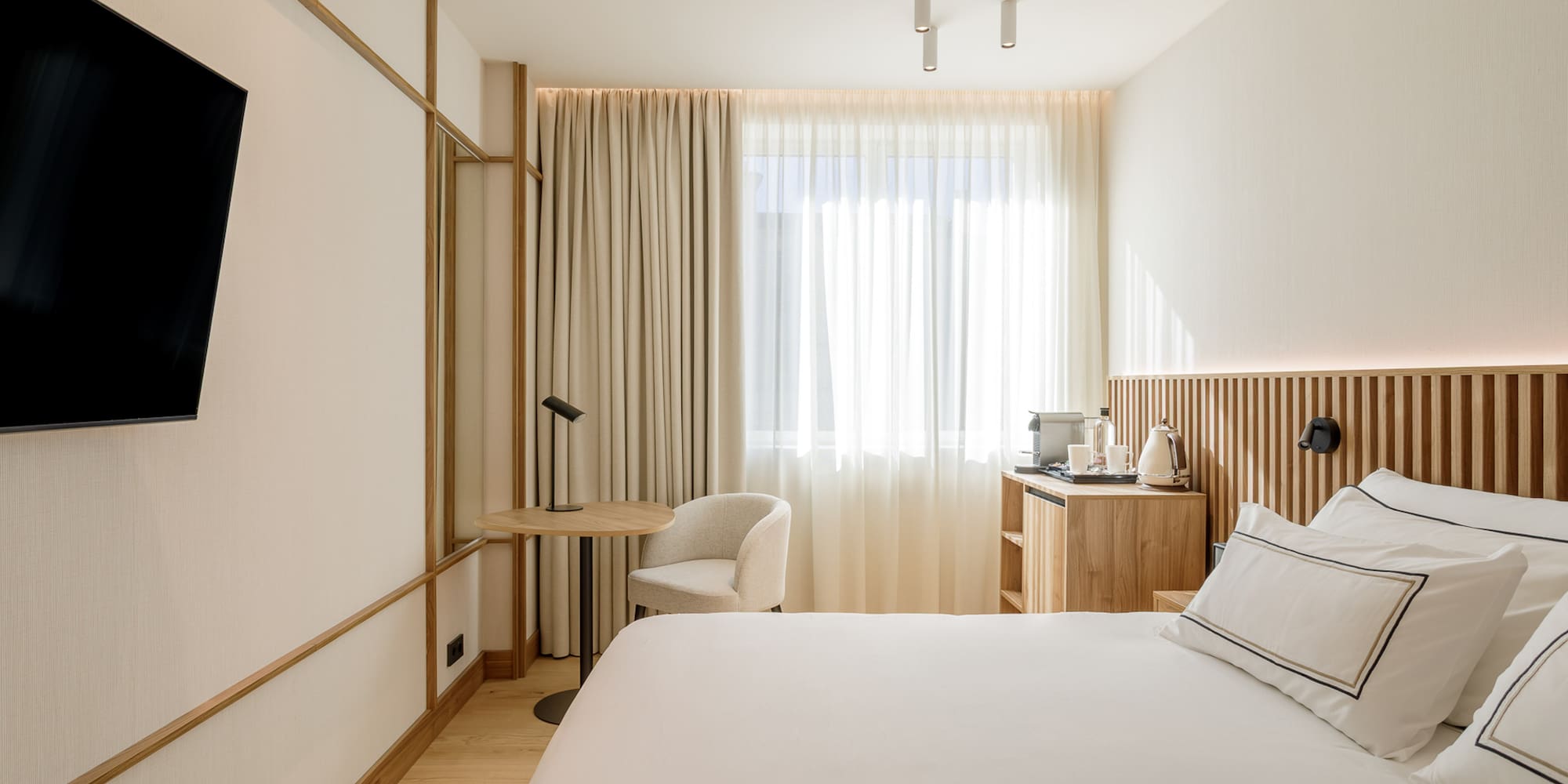 Minimalist hotel bedroom with white bed, wood accents, TV, and bright window.