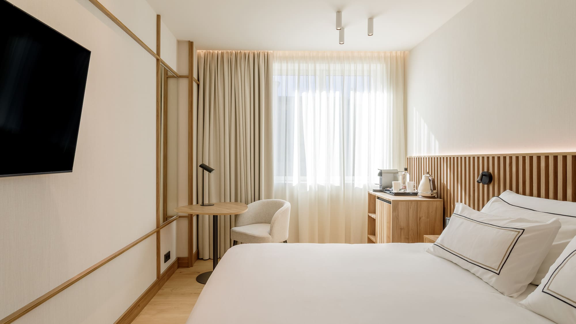Modern hotel room with white bed, wooden headboard, wall TV, and sunny window.