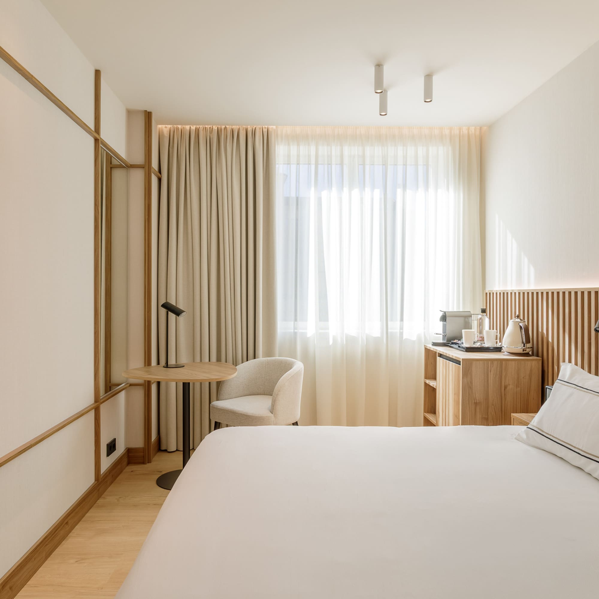 Minimalist hotel bedroom with white bed, wood accents, TV, and bright window.
