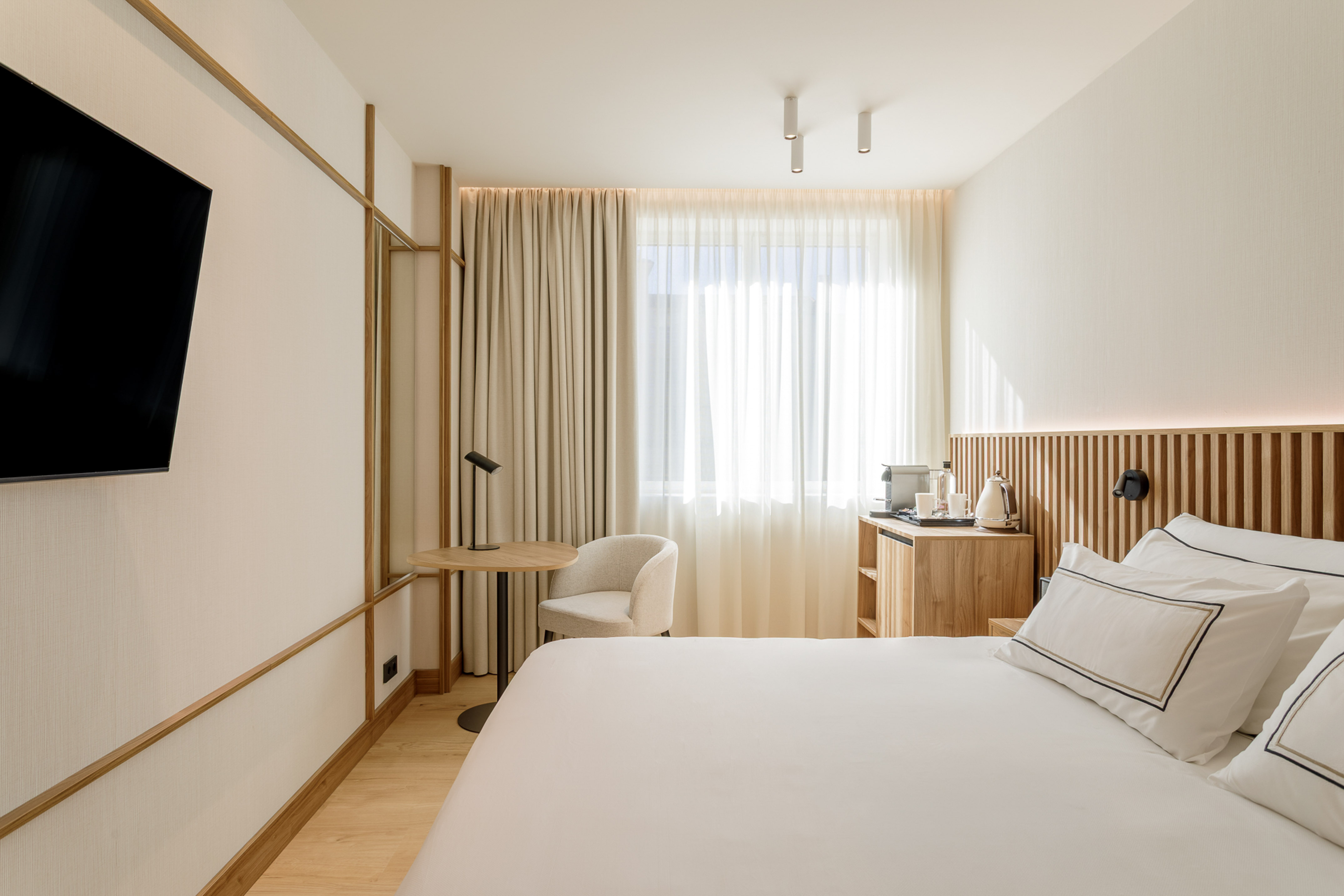 Modern hotel room with white bed, wooden headboard, wall TV, and sunny window.