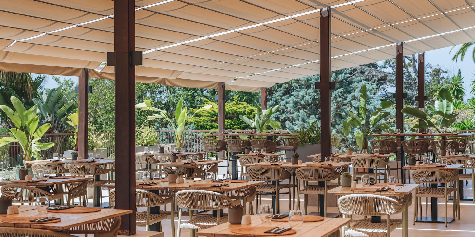 Shaded outdoor restaurant dining area with modern tables and lush greenery.