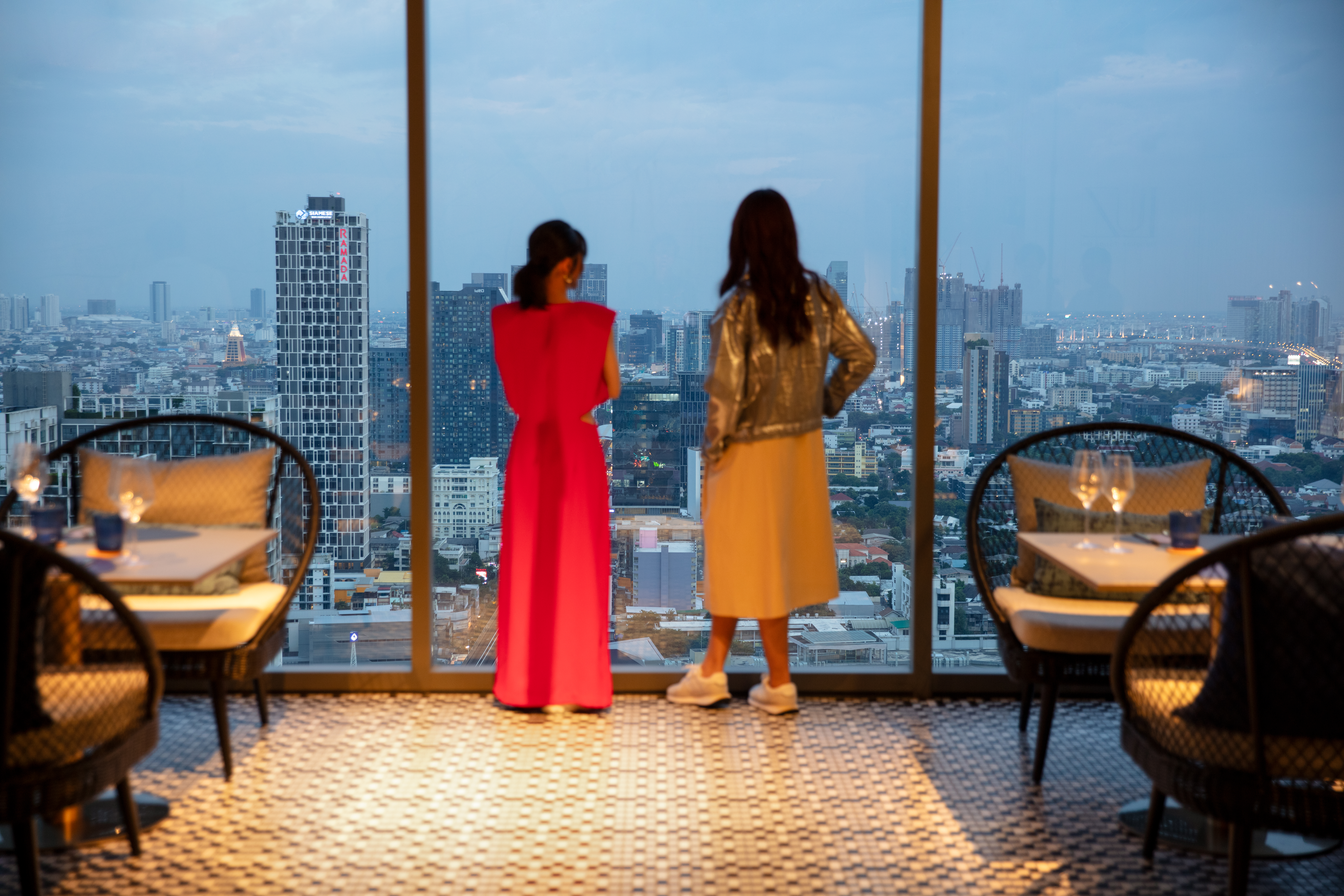 Two women viewing city skyline from high-rise restaurant at dusk.