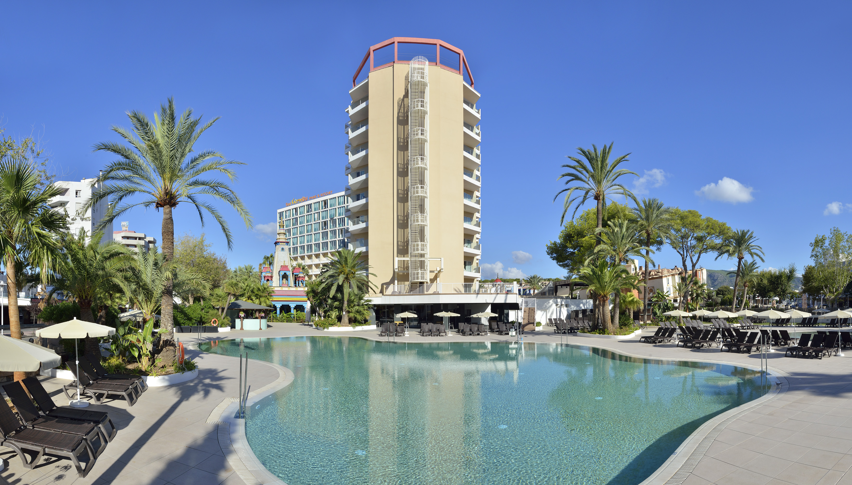 Modern hotel with large outdoor swimming pool, sun loungers, and palm trees.