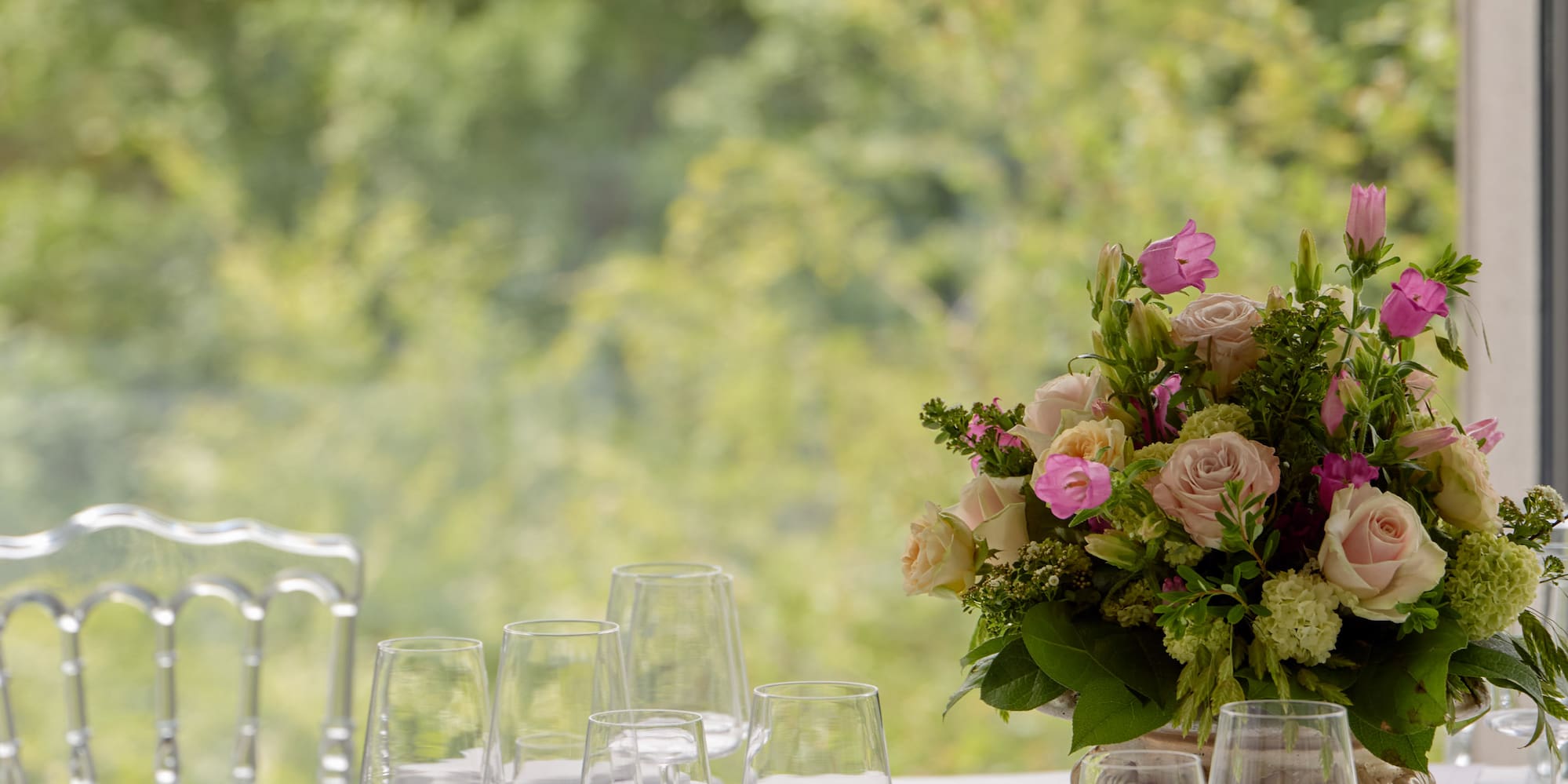 Elegant table set for wedding with floral centerpiece, clear chairs, outdoor view.
