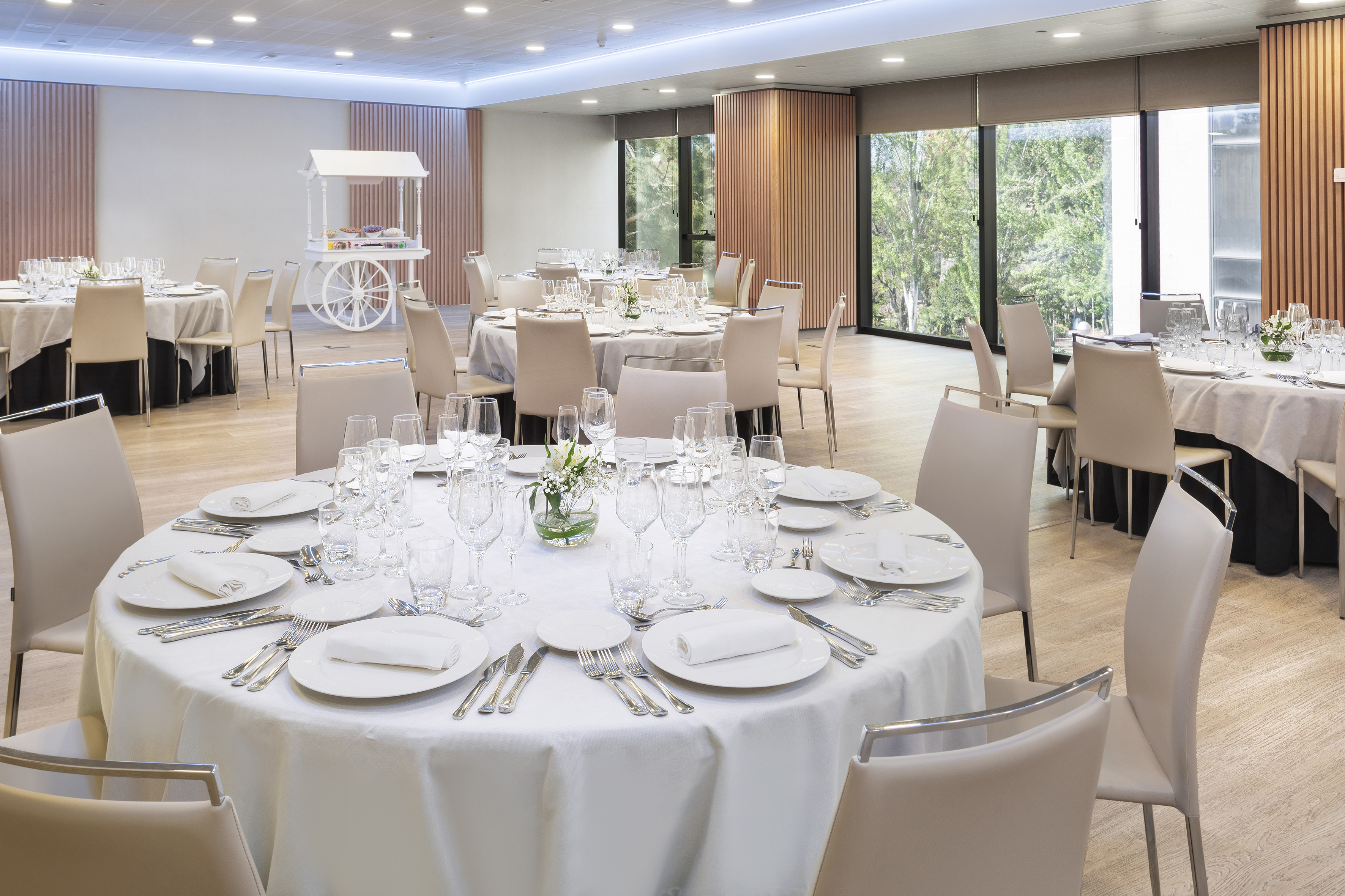Bright event room with round dining tables, beige chairs, and a dessert cart.