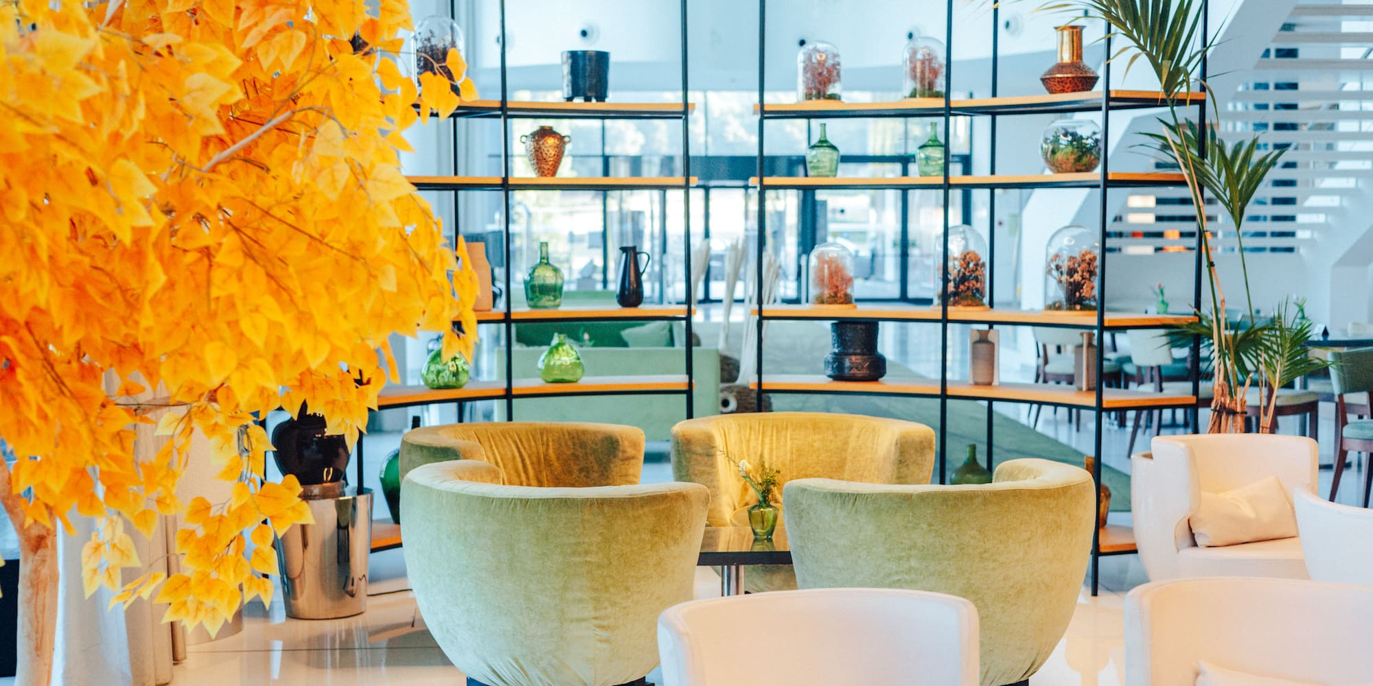 Modern lounge: bright yellow tree, green armchairs, white seating, and shelves.