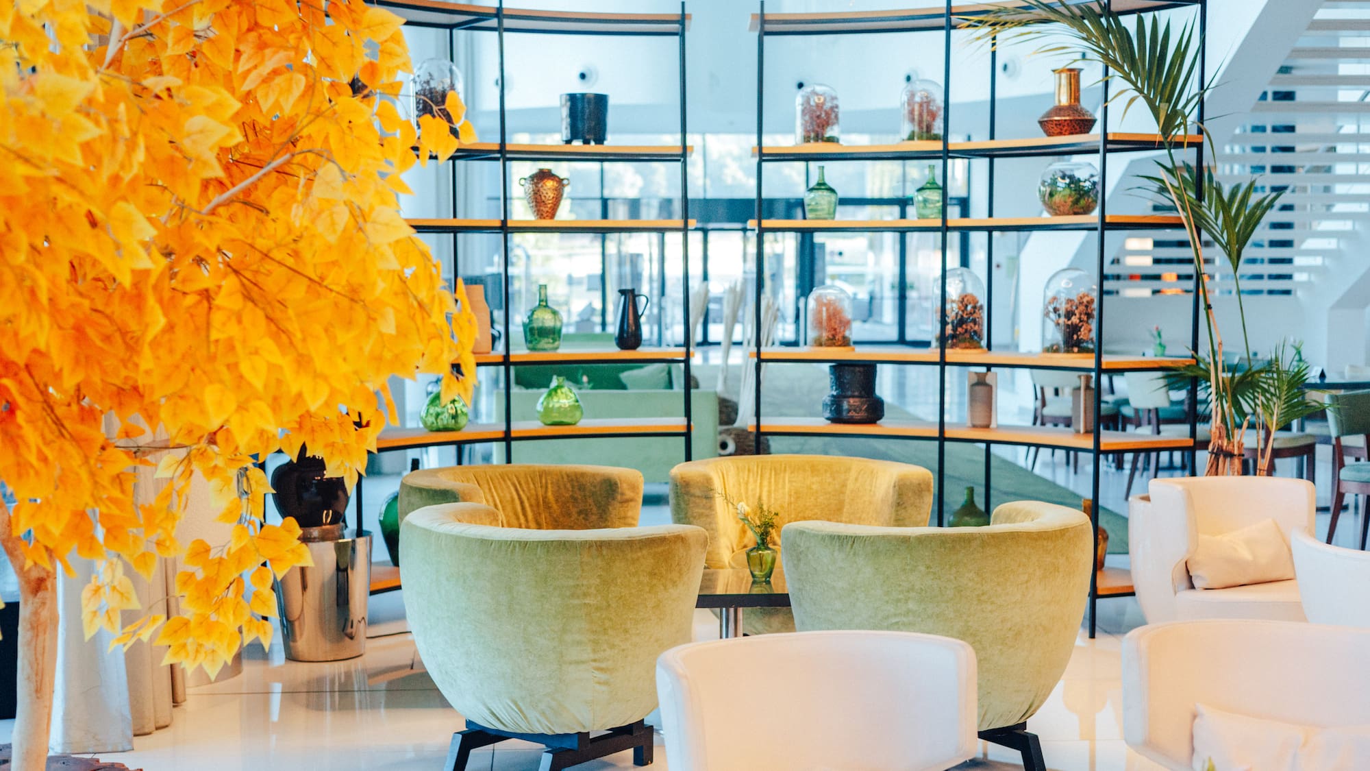 Modern lounge: bright yellow tree, green armchairs, white seating, and shelves.