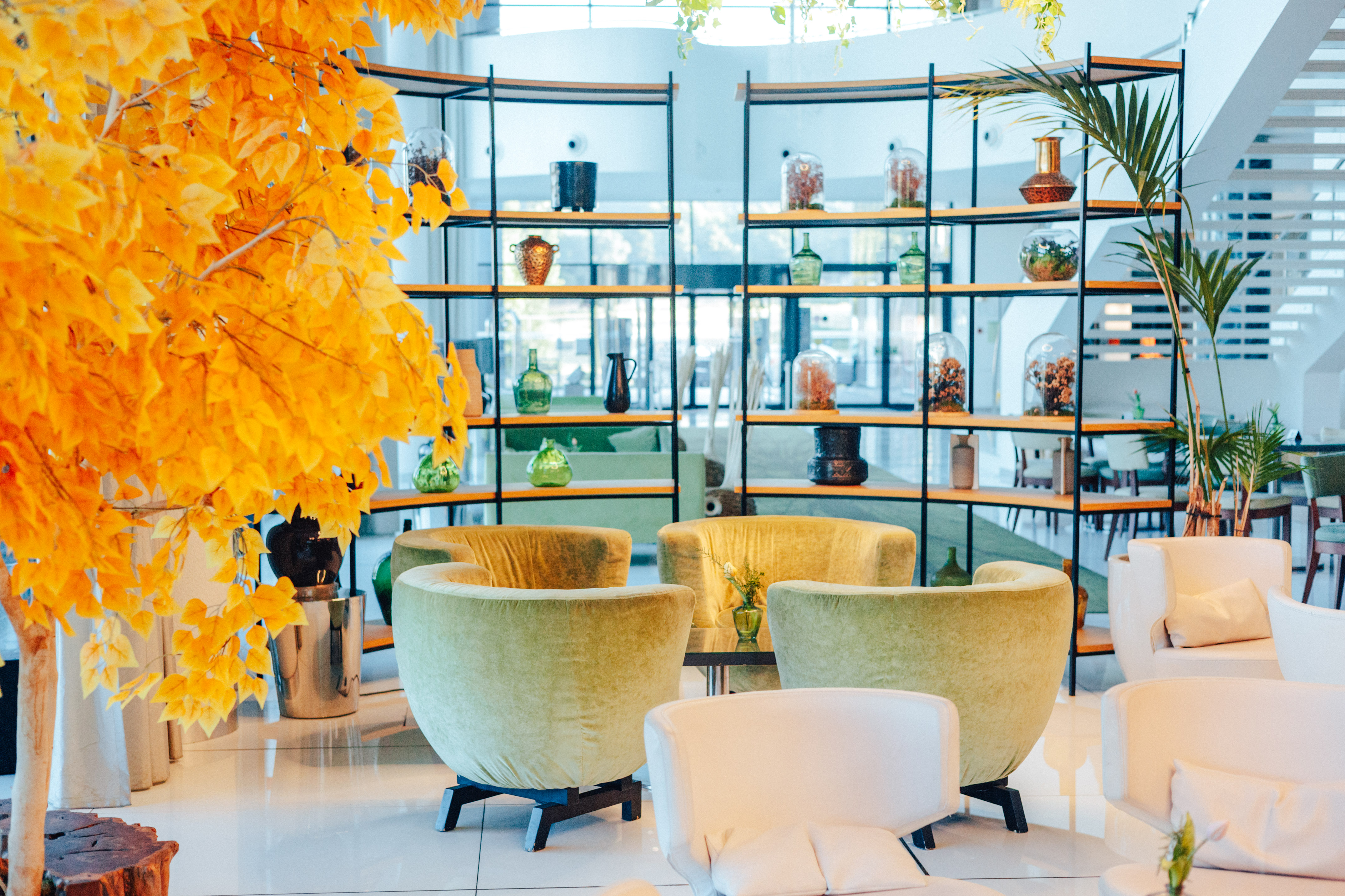 Modern lounge: bright yellow tree, green armchairs, white seating, and shelves.