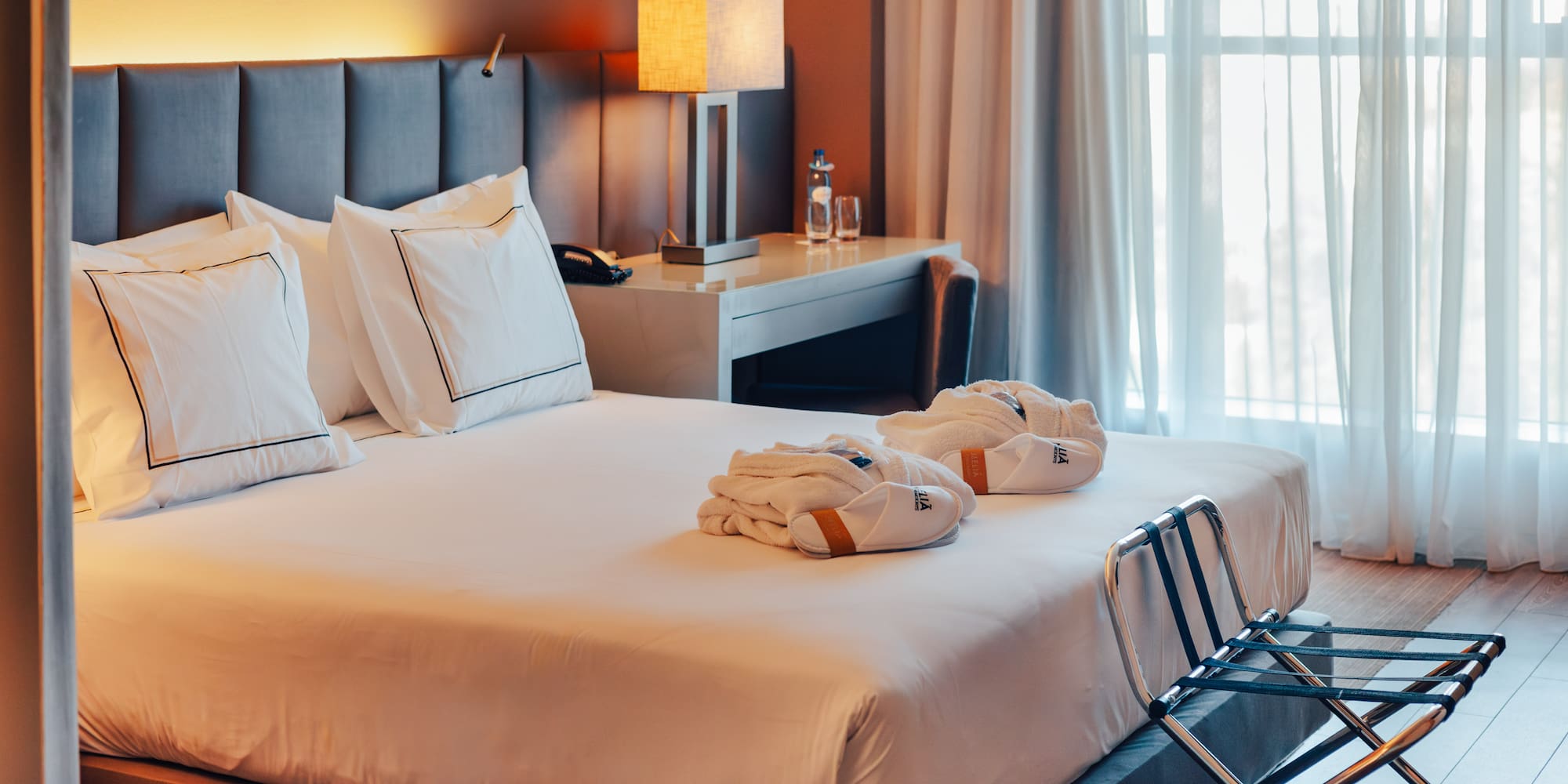 Hotel bedroom with large bed, gray headboard, bathrobes, and sunlit window.