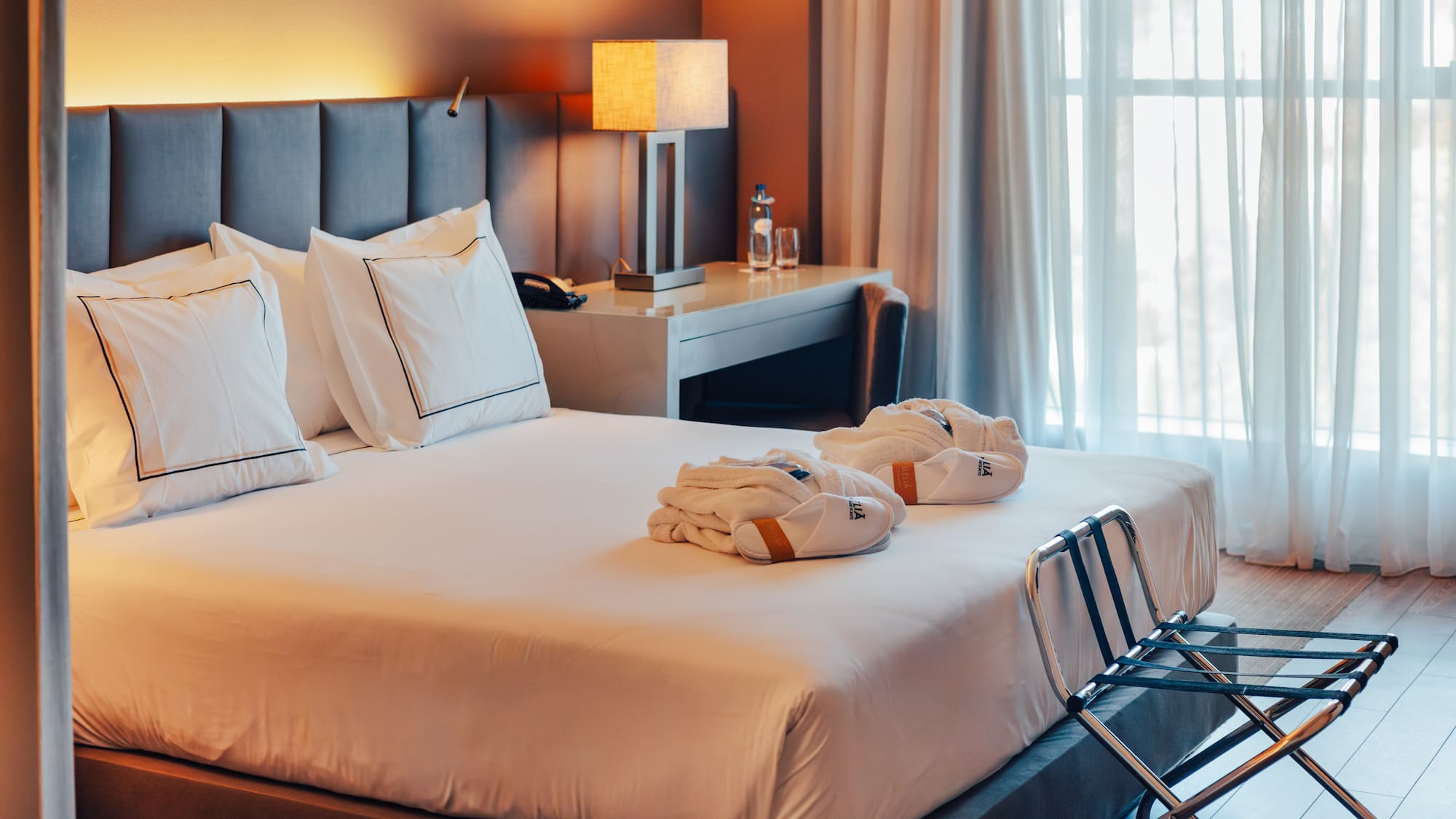 Hotel bedroom with large bed, gray headboard, bathrobes, and sunlit window.