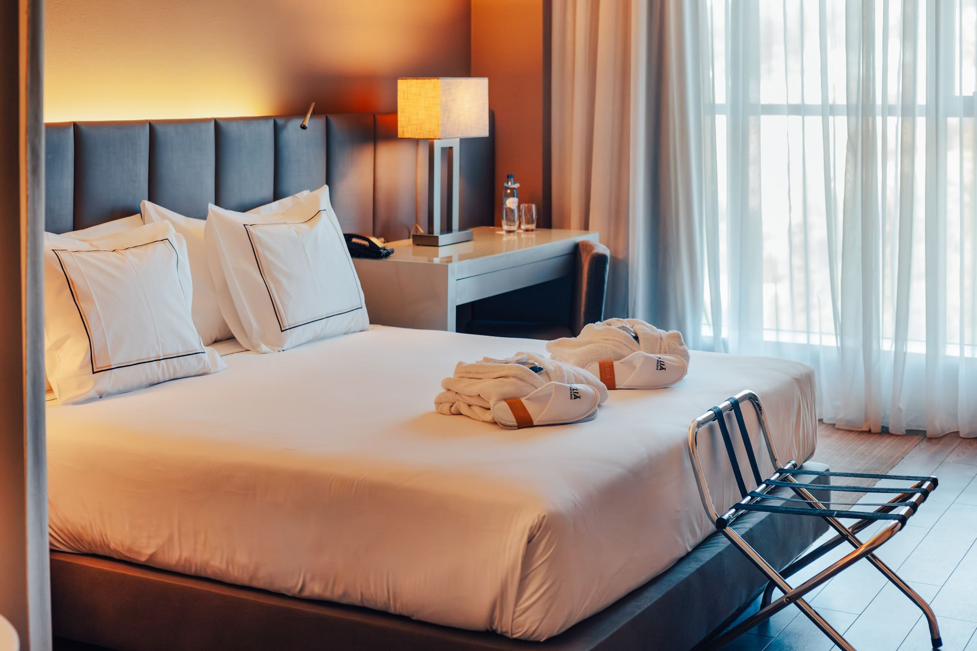 Hotel bedroom with large bed, gray headboard, bathrobes, and sunlit window.