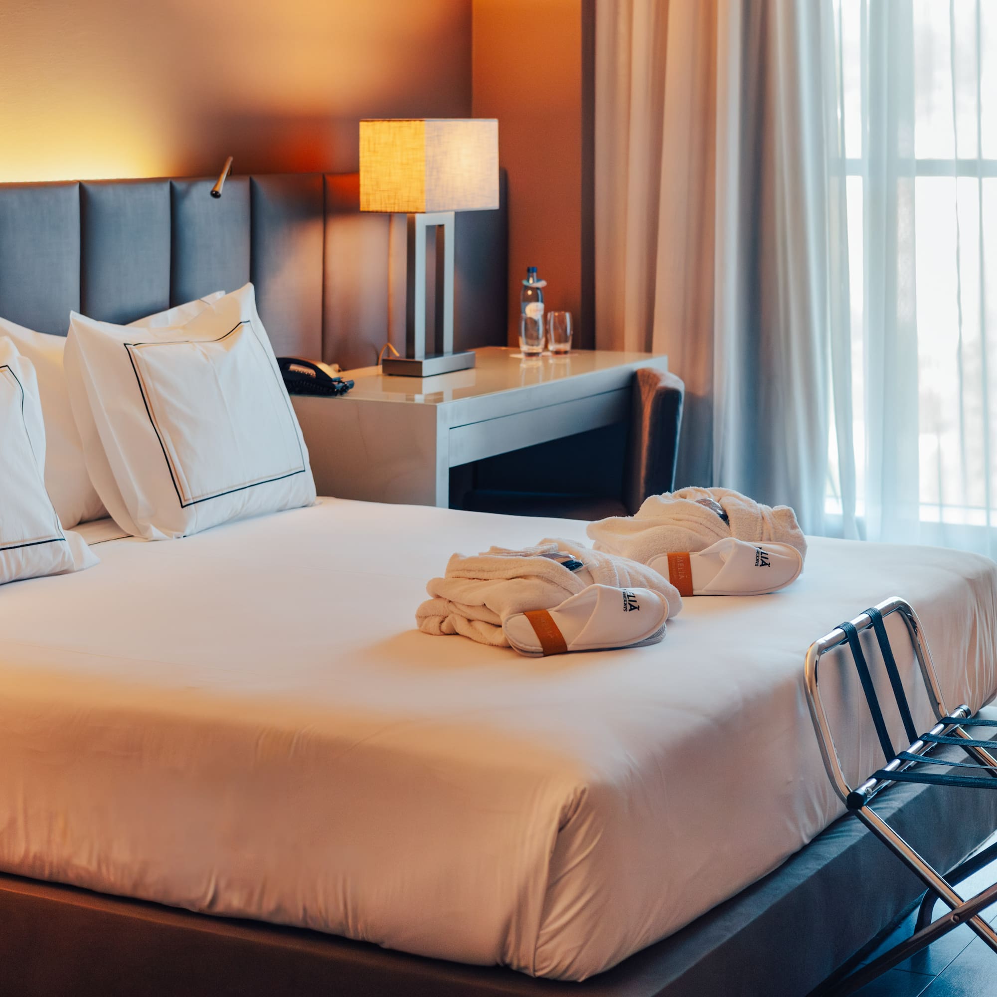 Hotel bedroom with large bed, gray headboard, bathrobes, and sunlit window.
