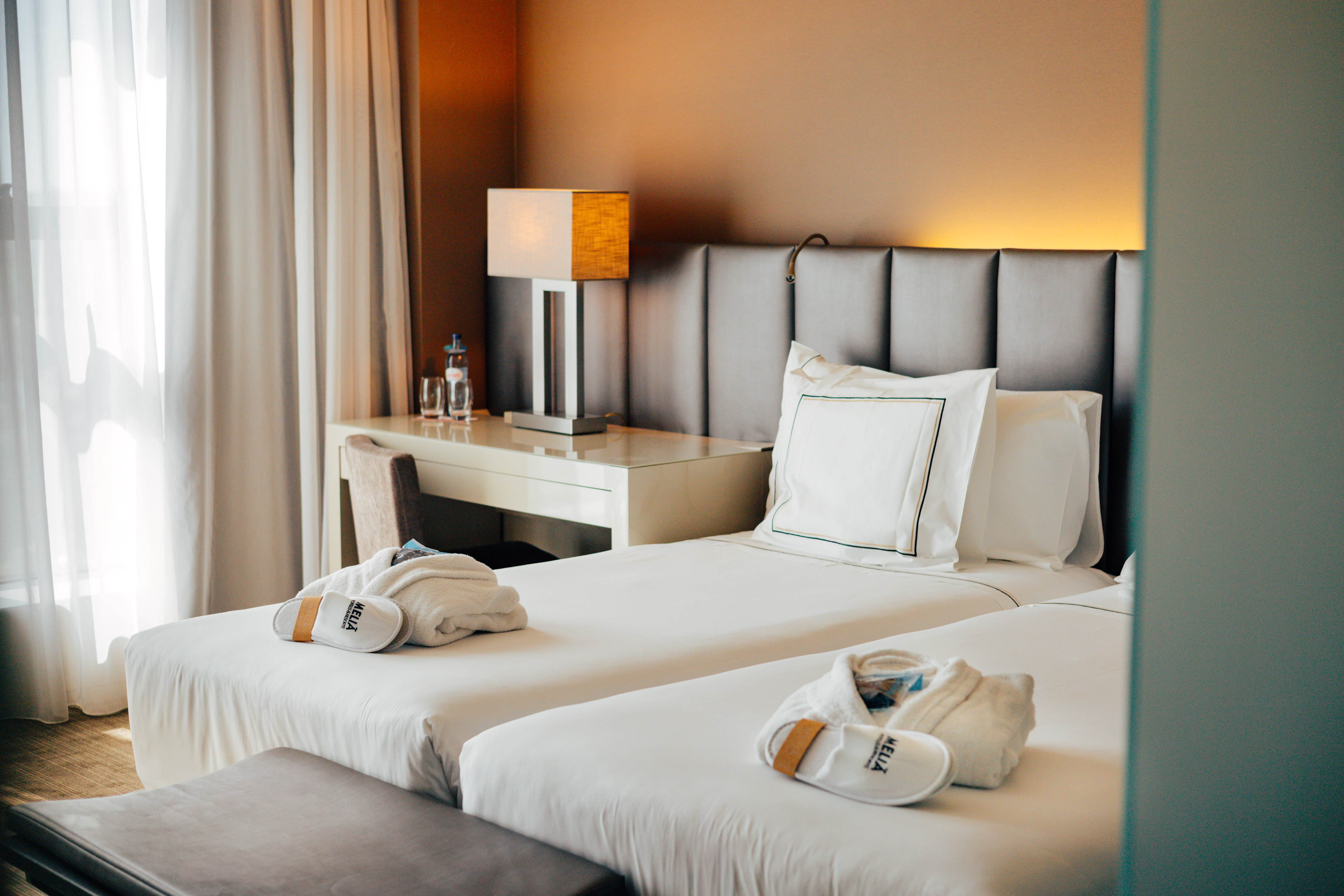 Hotel twin room with white bedding, Meliá robes, slippers, and desk lamp.
