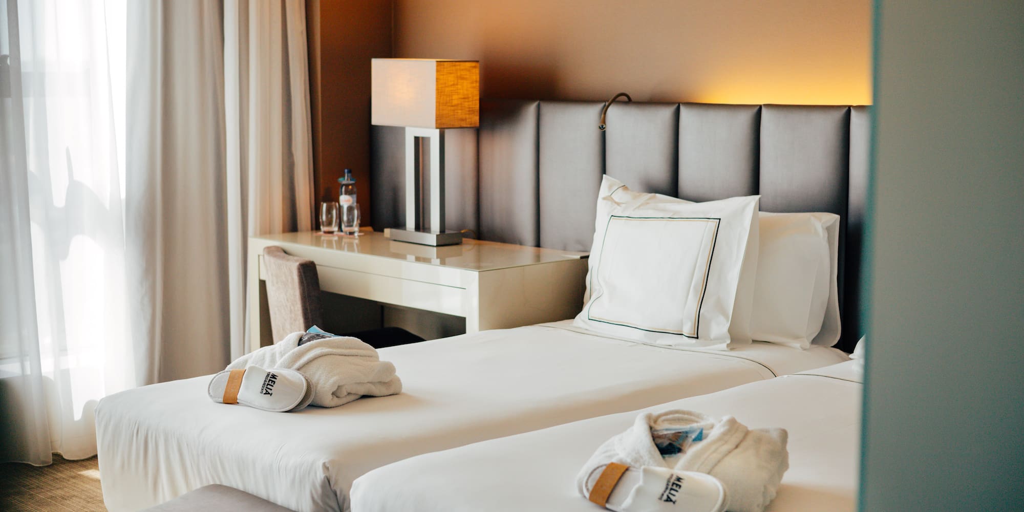 Hotel twin room with white bedding, Meliá robes, slippers, and desk lamp.