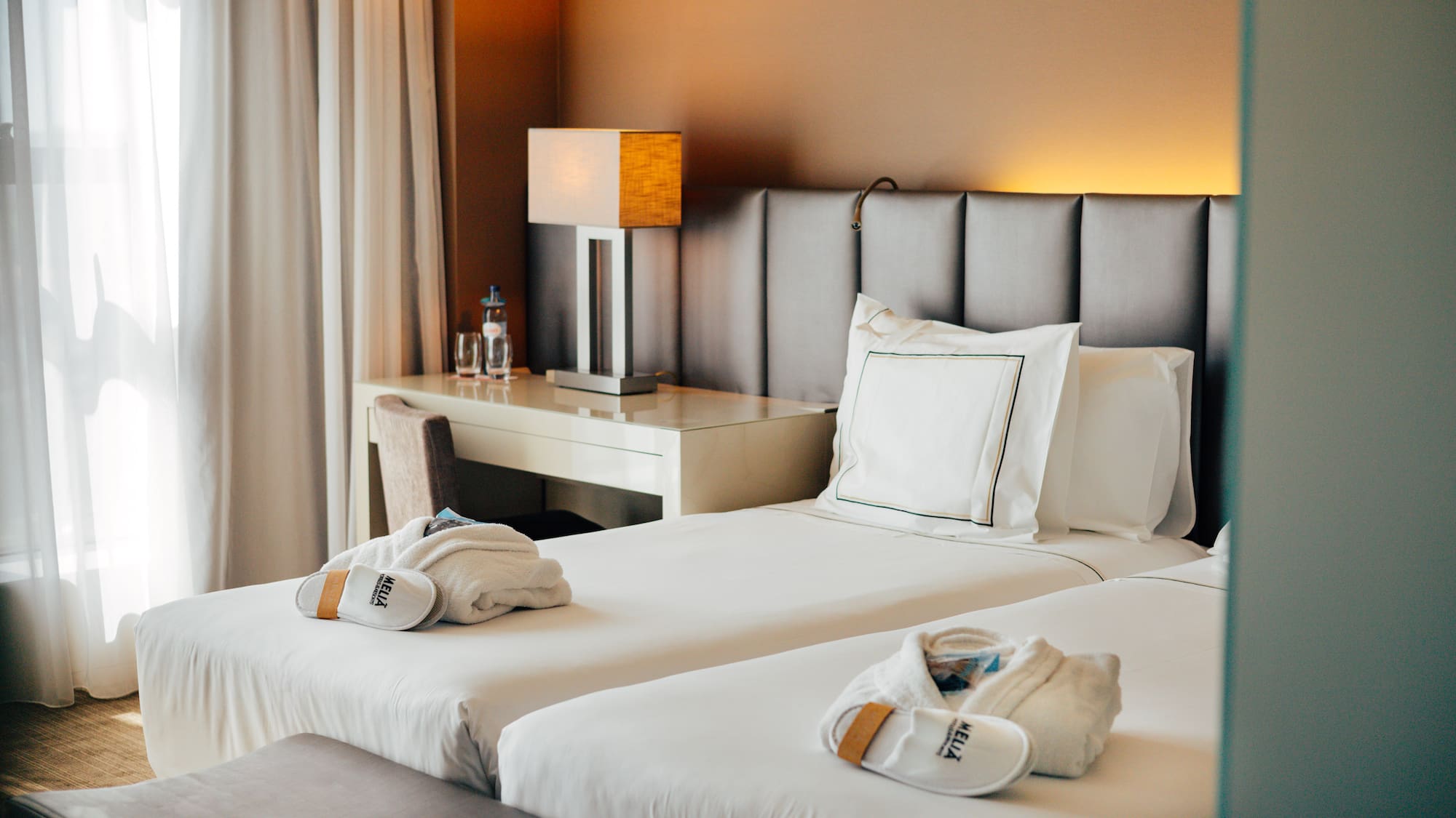 Hotel twin room with white bedding, Meliá robes, slippers, and desk lamp.