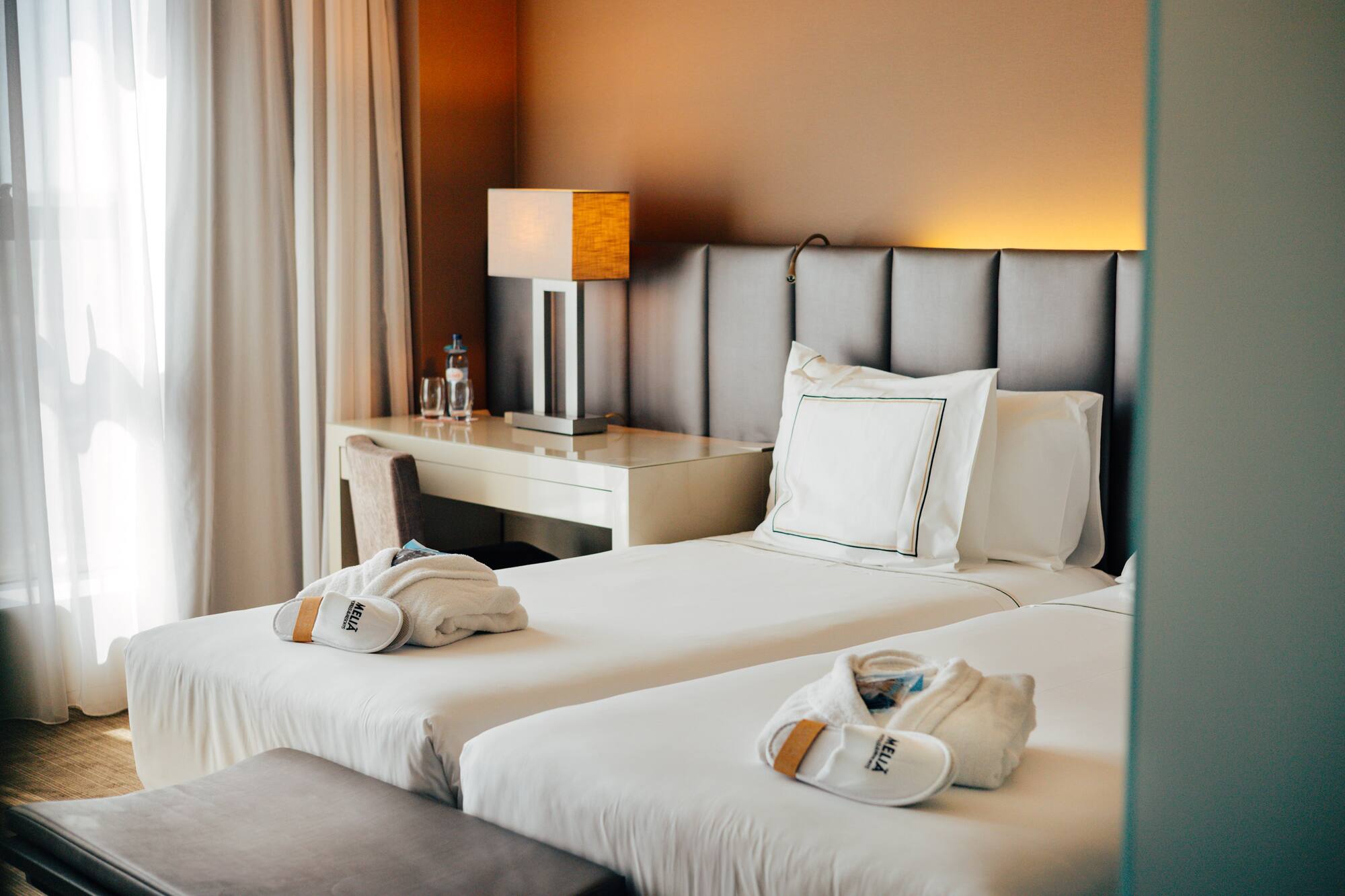 Hotel twin room with white bedding, Meliá robes, slippers, and desk lamp.