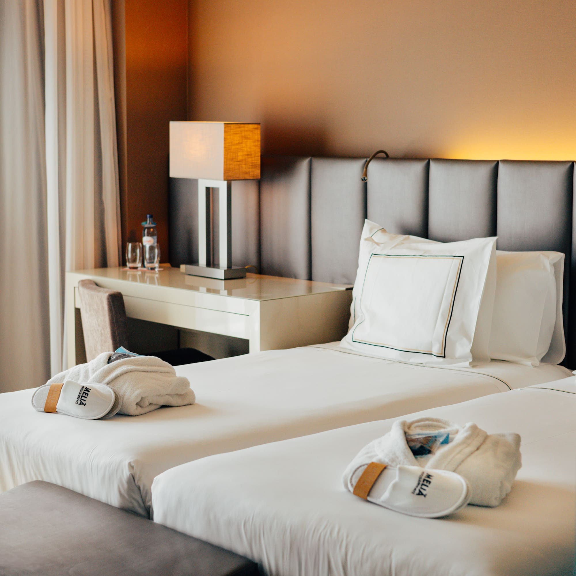 Hotel twin room with white bedding, Meliá robes, slippers, and desk lamp.