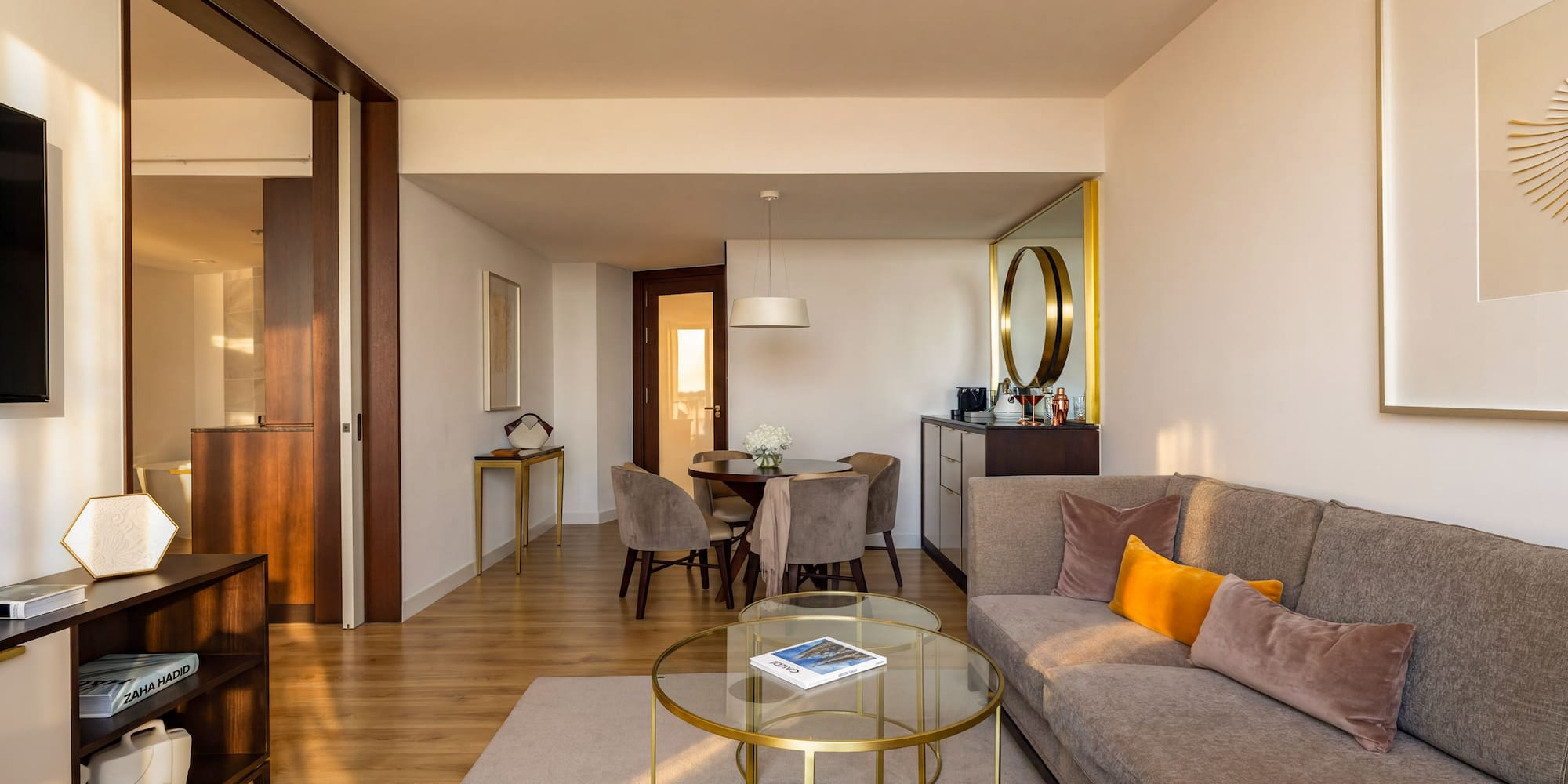 Modern hotel suite living room with dining area, gray sofa, and wood floors.
