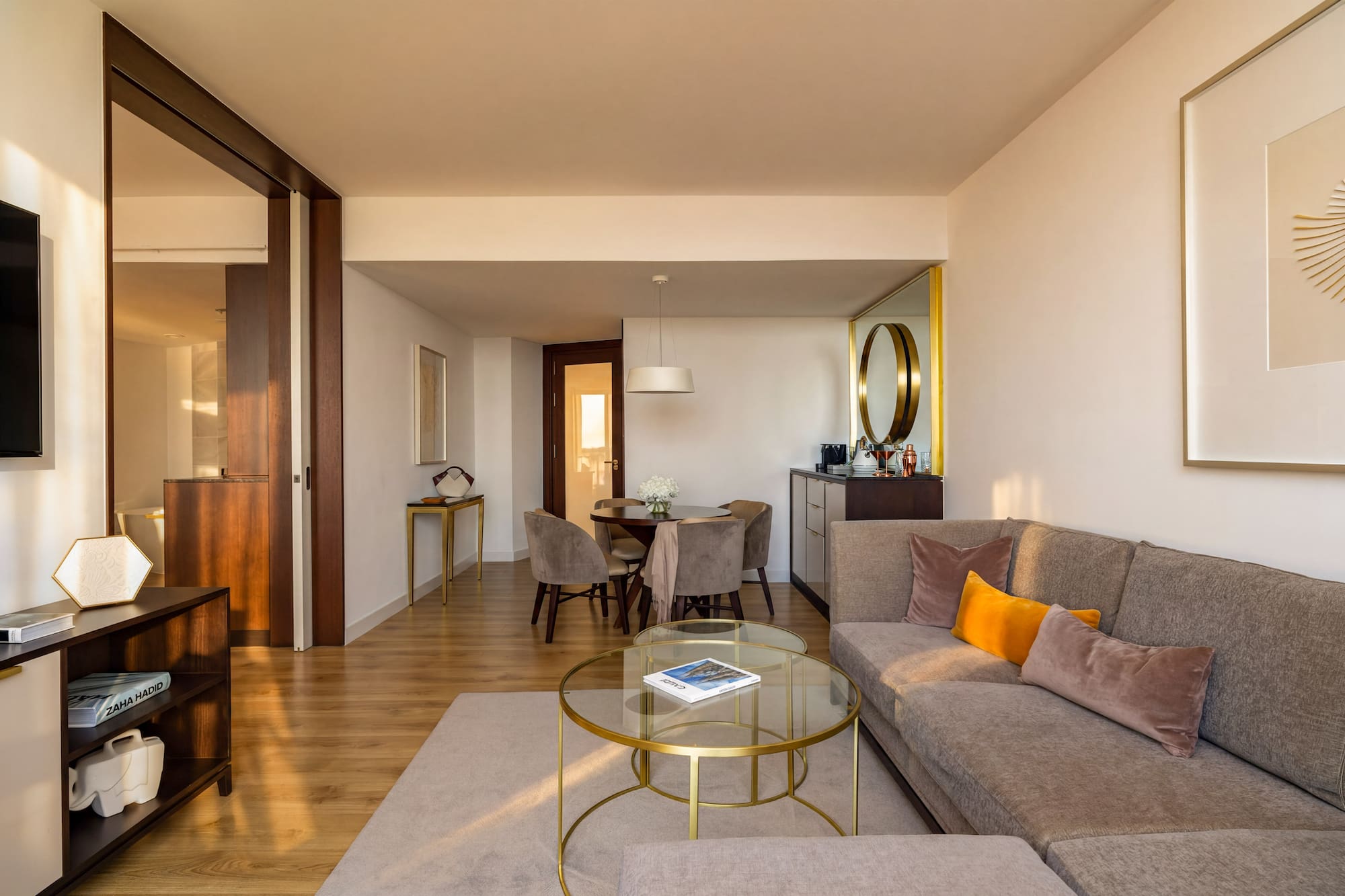 Modern hotel suite living room with dining area, gray sofa, and wood floors.