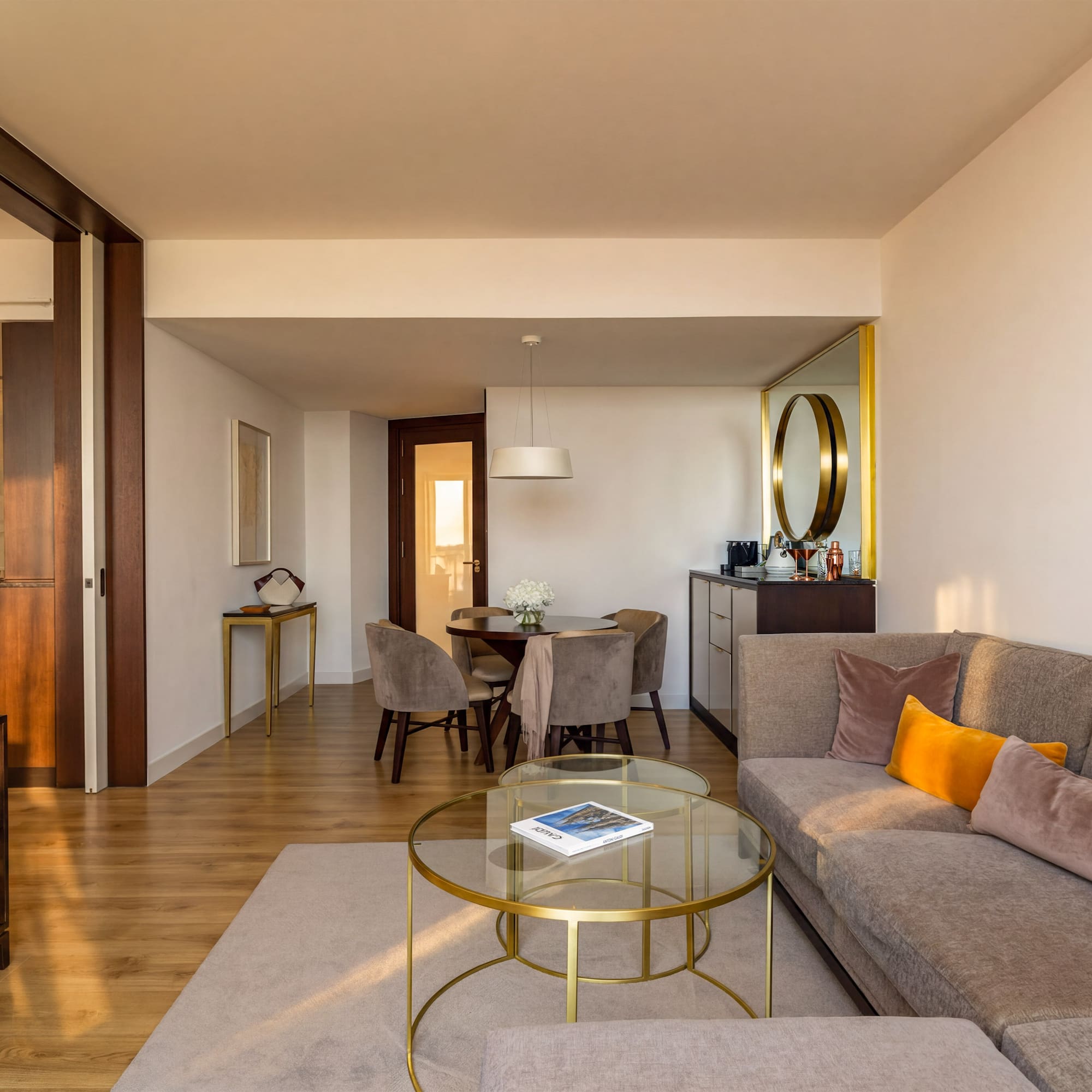 Modern hotel suite living room with dining area, gray sofa, and wood floors.