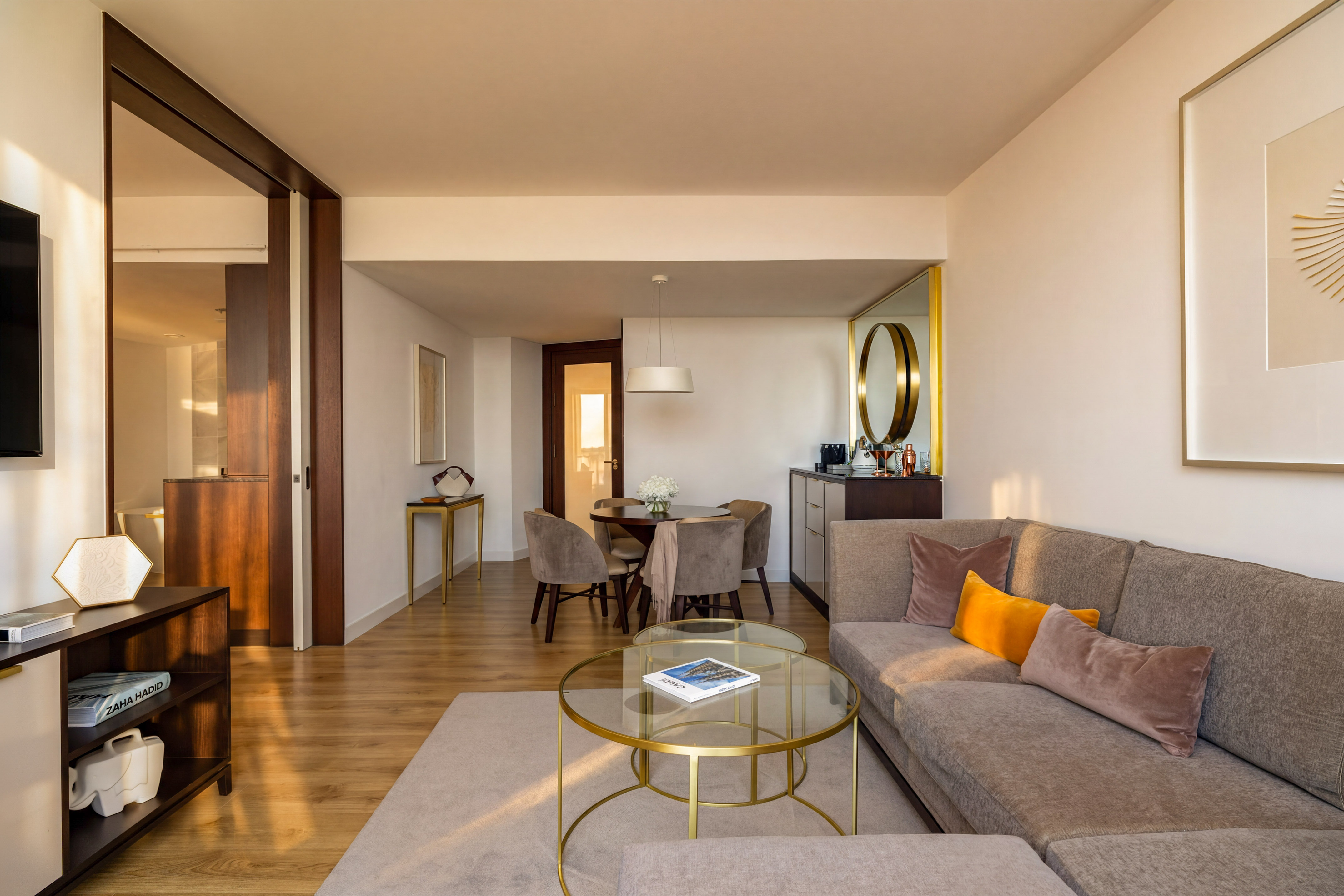 Modern hotel suite living room with dining area, gray sofa, and wood floors.