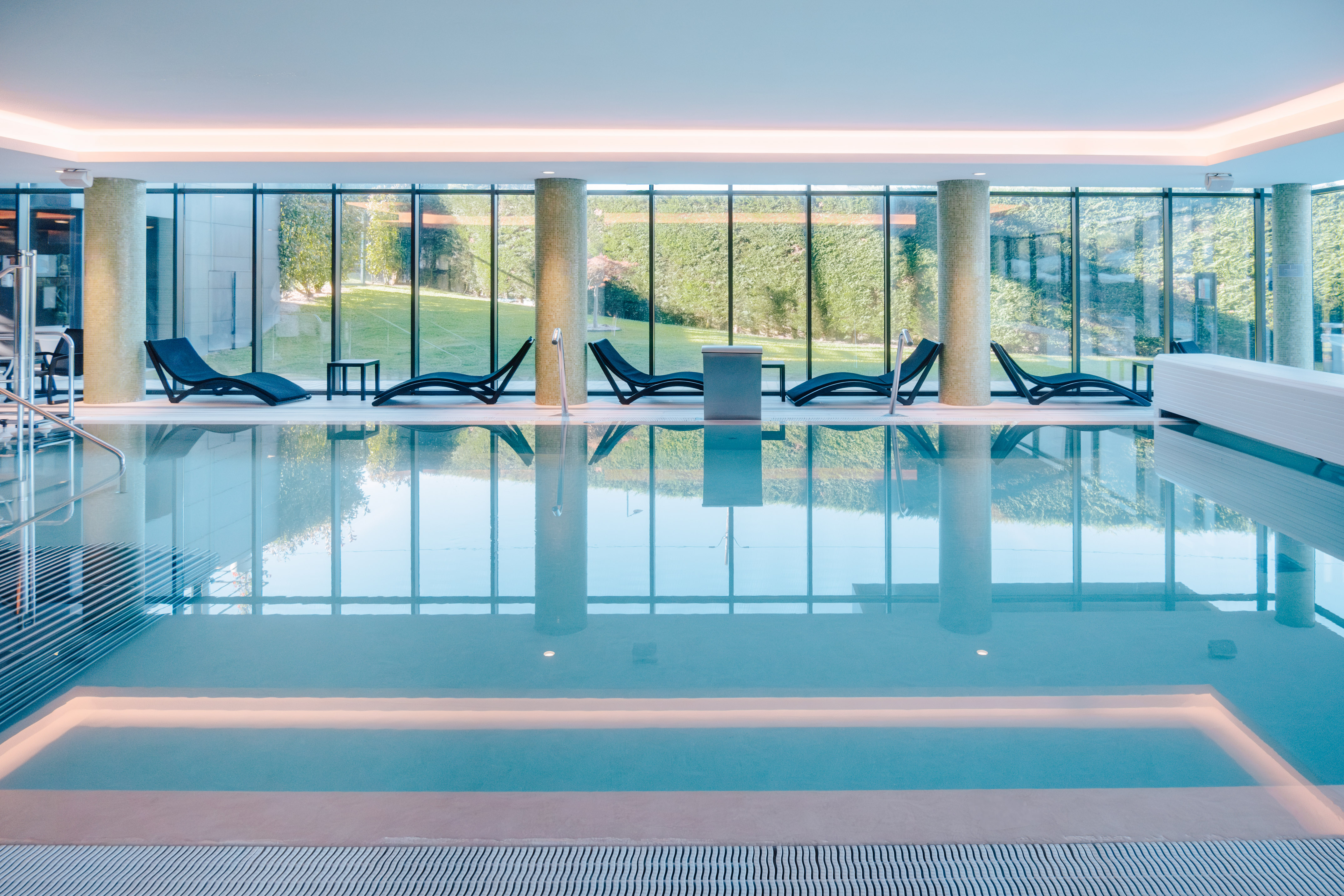 Modern indoor pool, lounge chairs, full-length windows, green outdoor view.