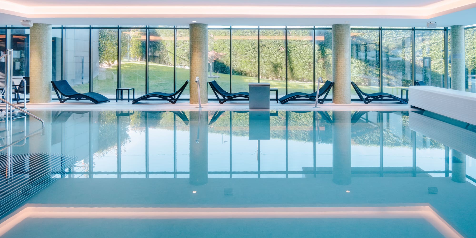 Modern indoor pool, lounge chairs, full-length windows, green outdoor view.
