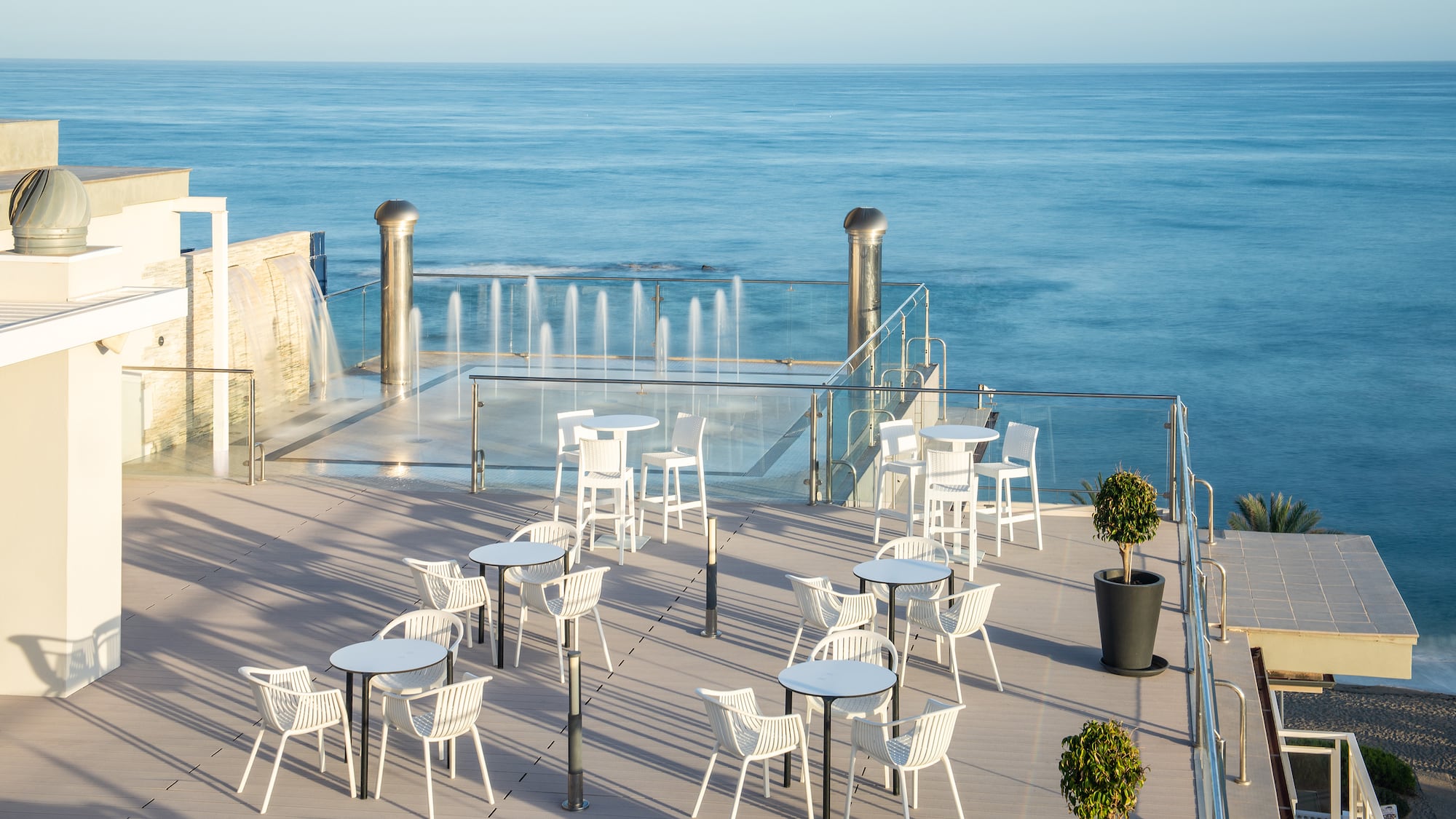 a patio with white tables and chairs and a fountain in front of the ocean