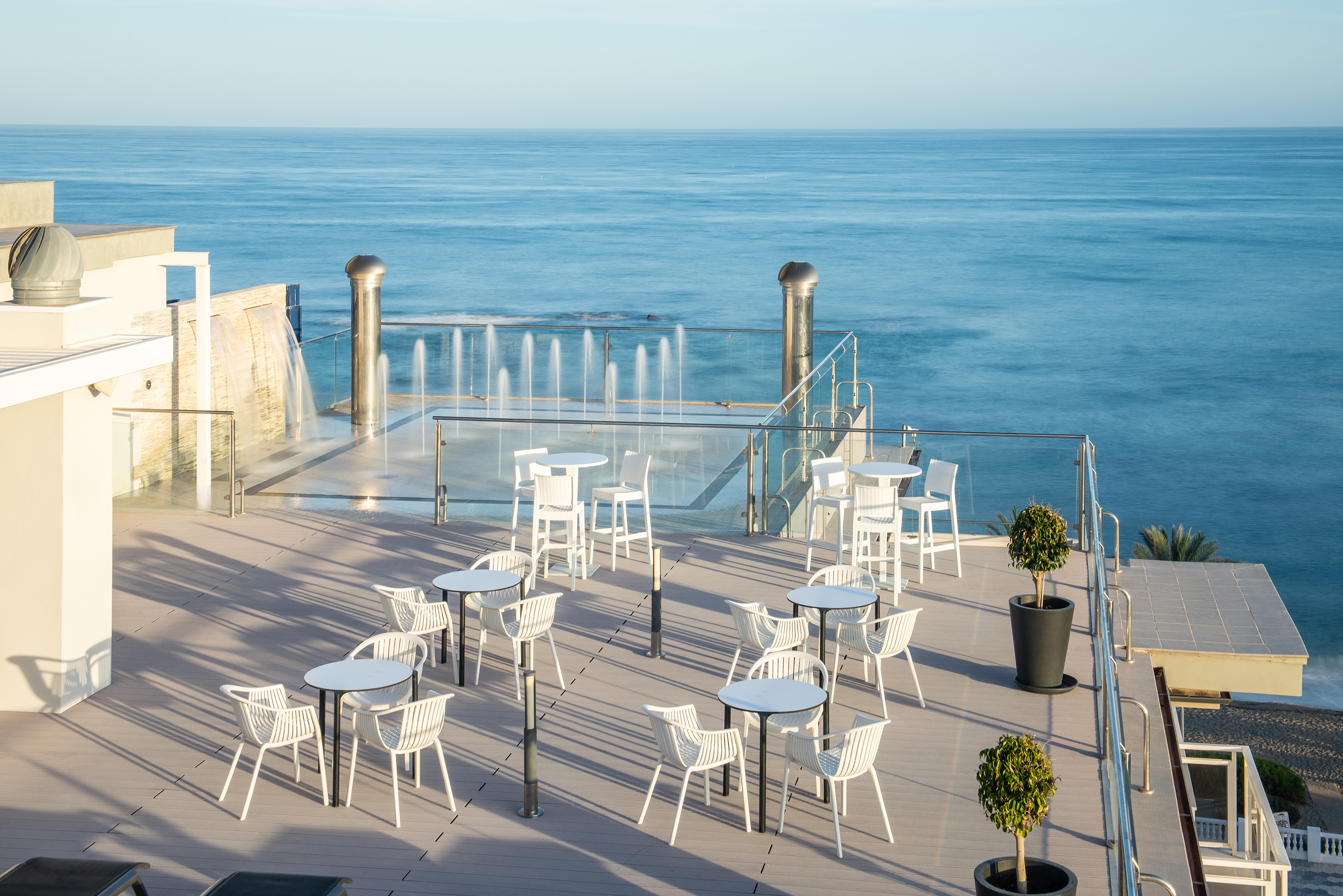 a patio with white tables and chairs and a fountain in front of the ocean