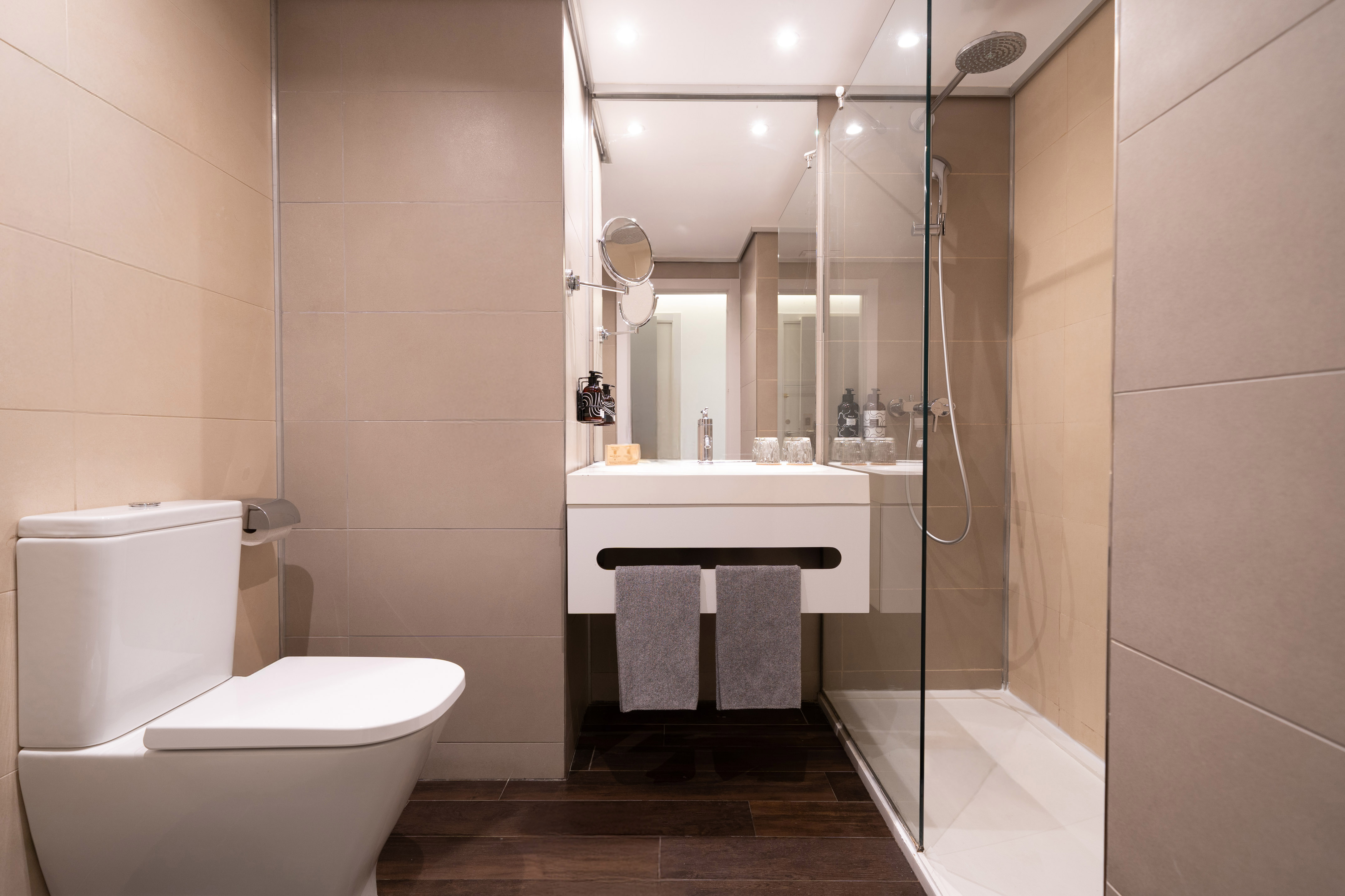 Modern hotel bathroom with beige tiles, white toilet, vanity, and glass shower.