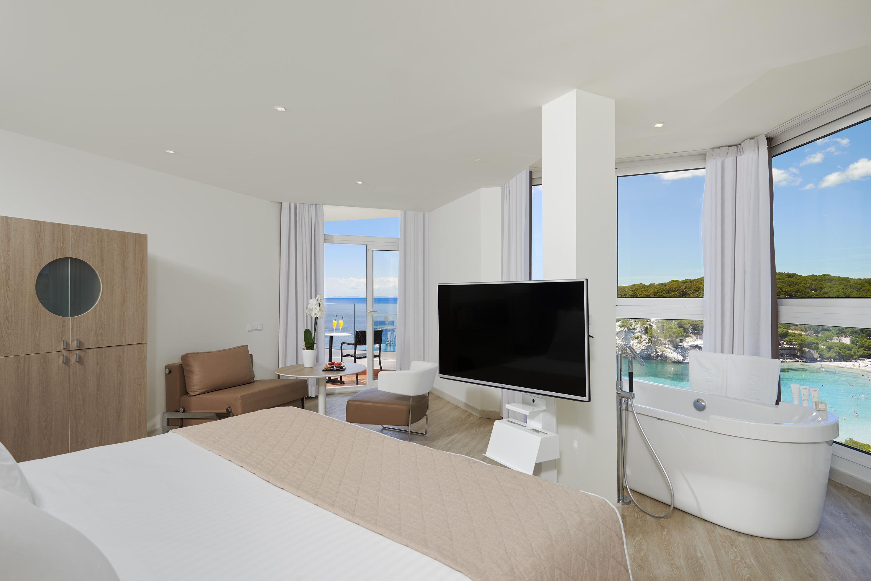Modern ocean view suite with balcony, bed, and bathtub by a sandy beach.