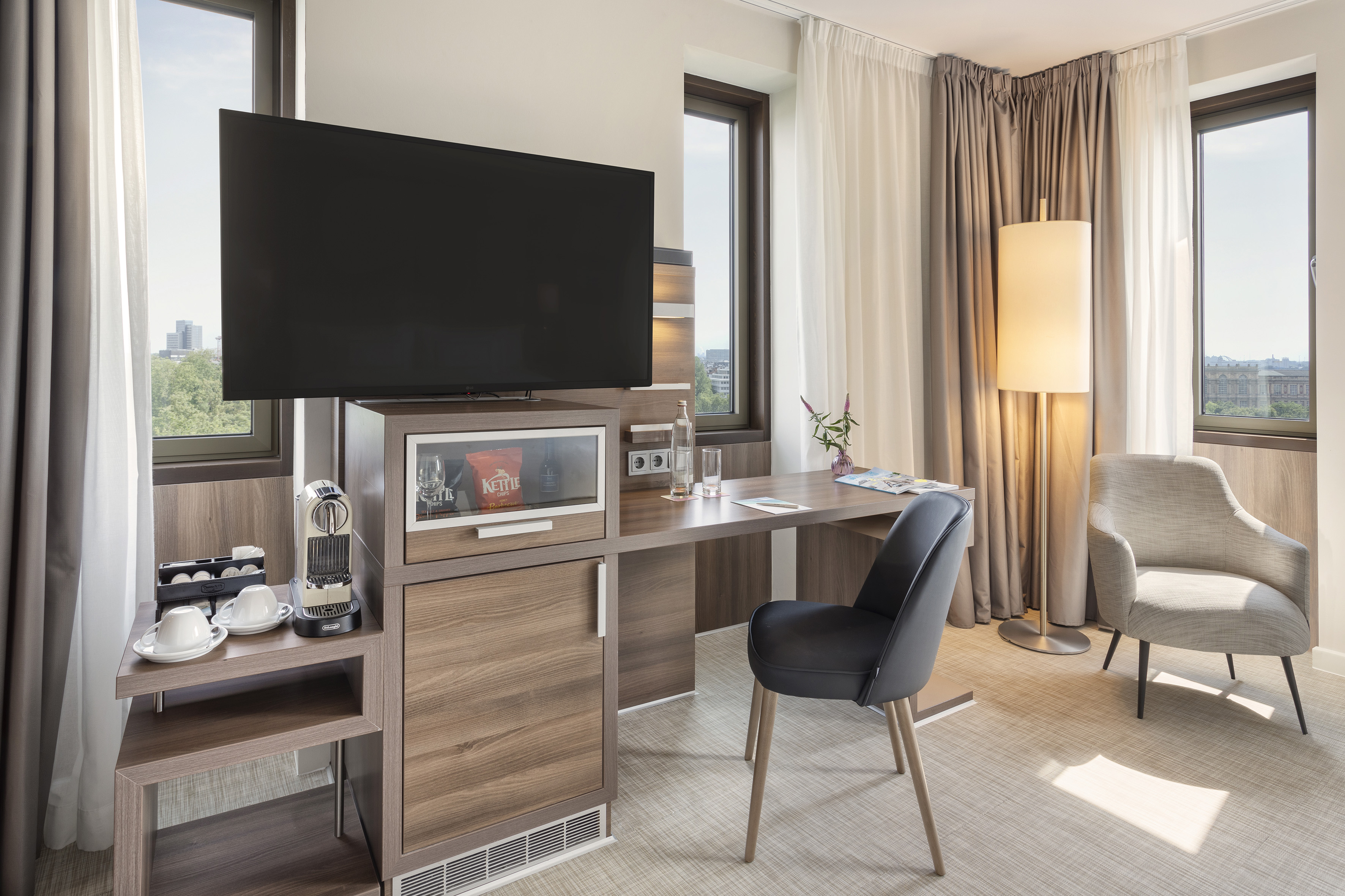 Modern hotel room with TV, desk, coffee machine, mini-bar, and armchair.