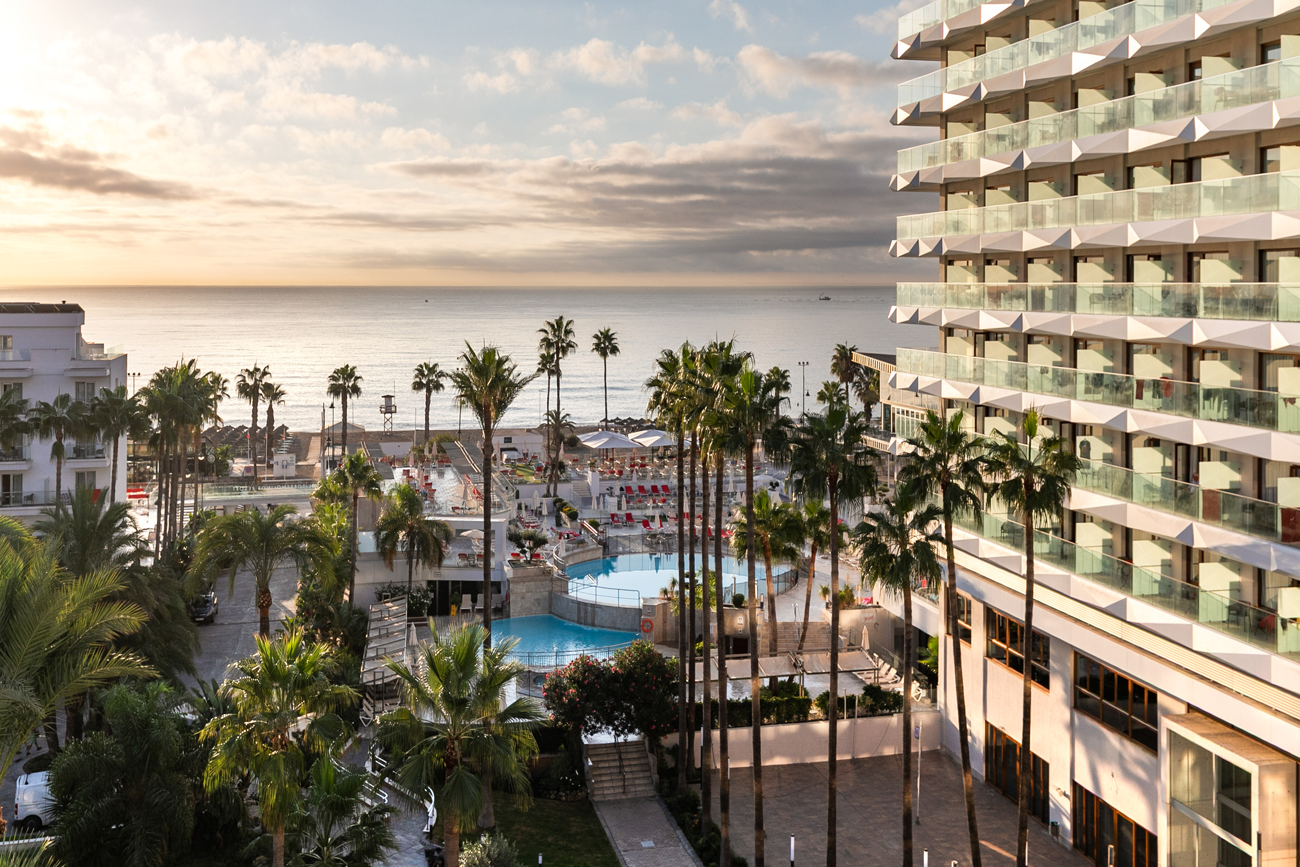 Beachfront resort hotel with pools, palm trees, and ocean view at sunrise.