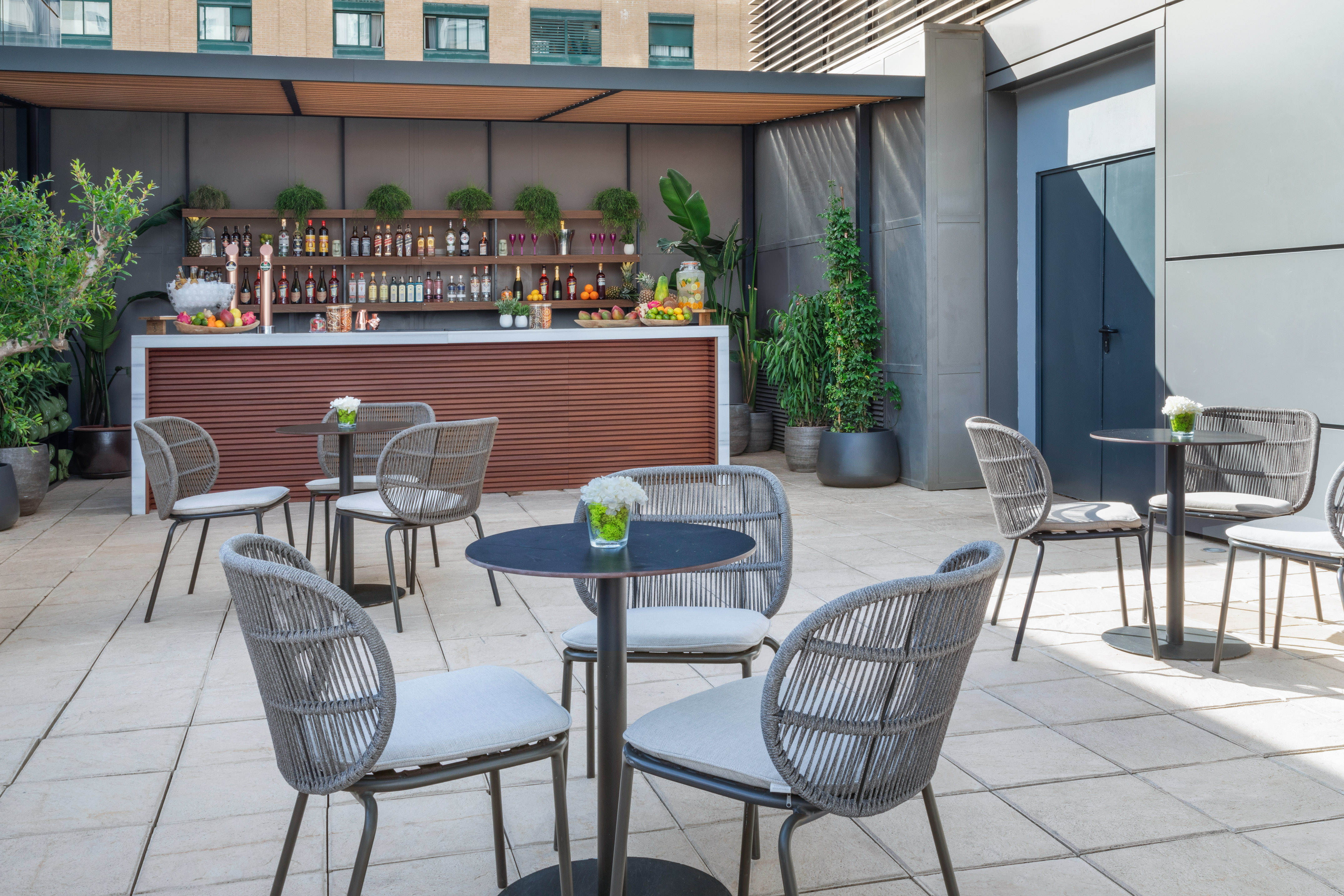 Outdoor patio bar with cocktail setup, gray chairs, and lush potted plants.