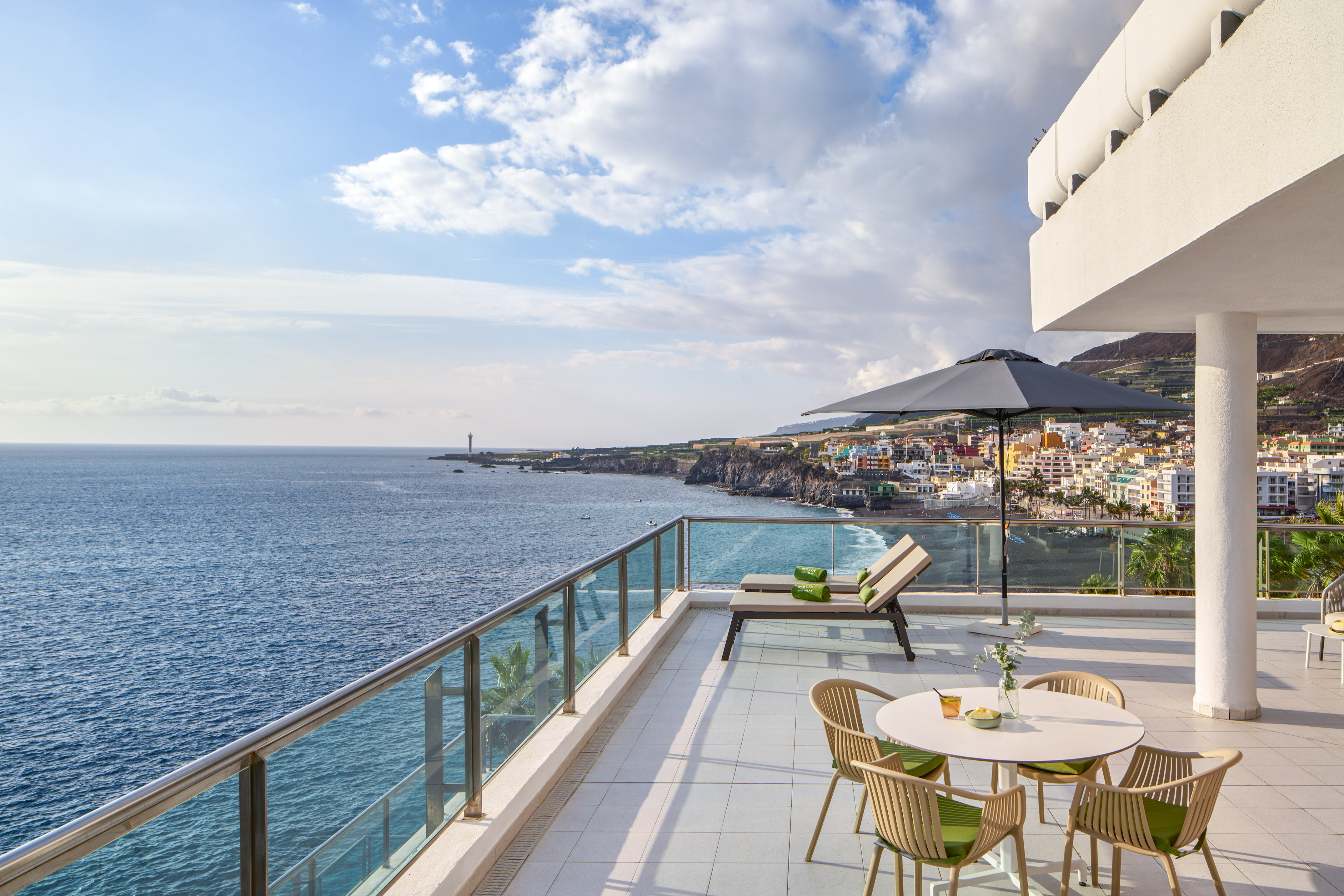 Sunny ocean view balcony with lounge chairs, coastal town, and lighthouse.