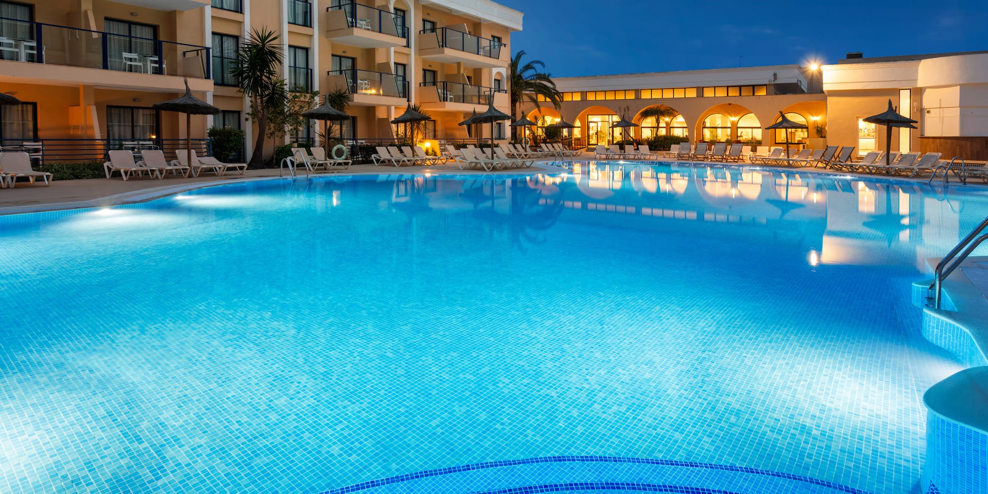 Evening view of illuminated resort pool with hotel building and palm trees.