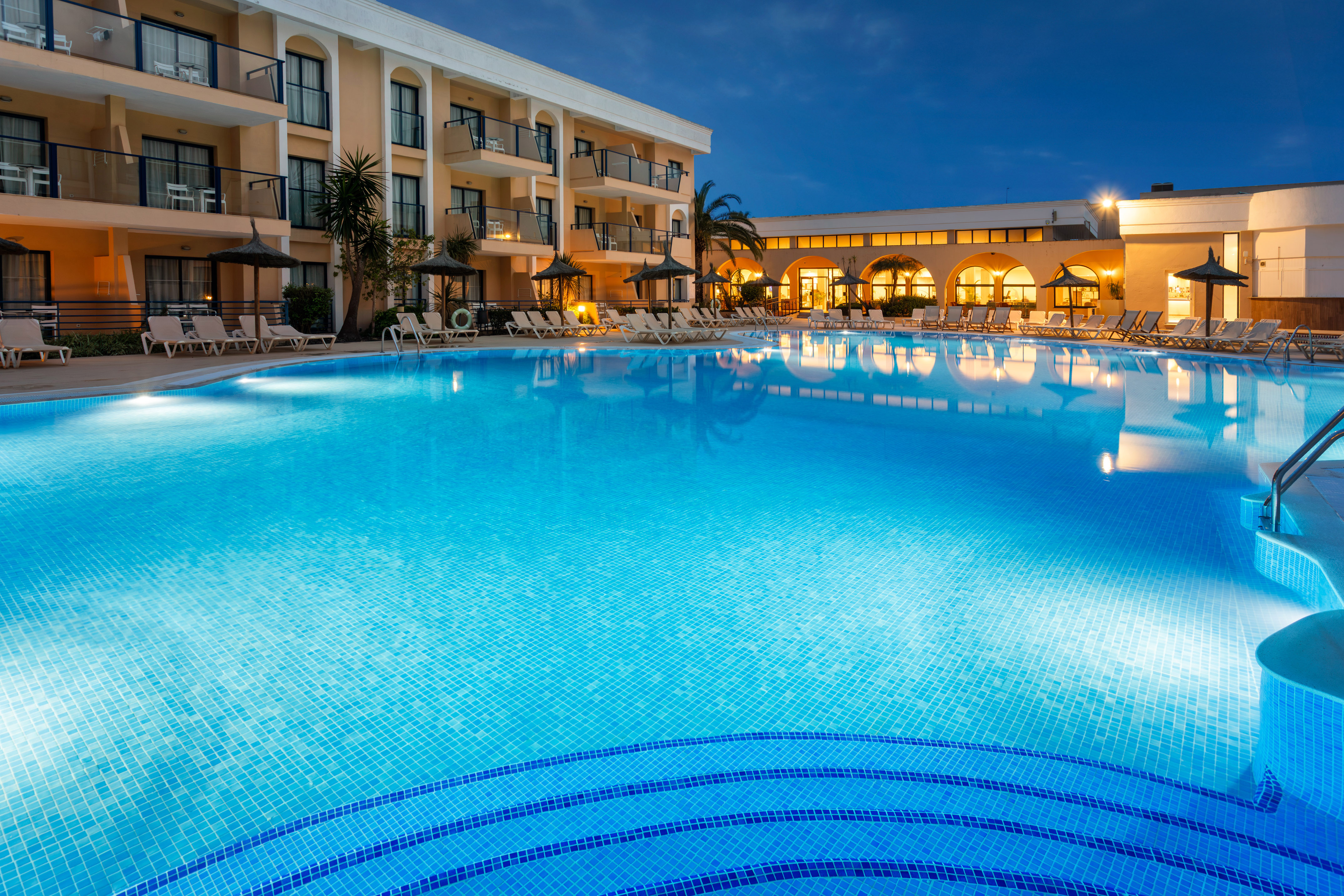 Evening view of illuminated resort pool with hotel building and palm trees.