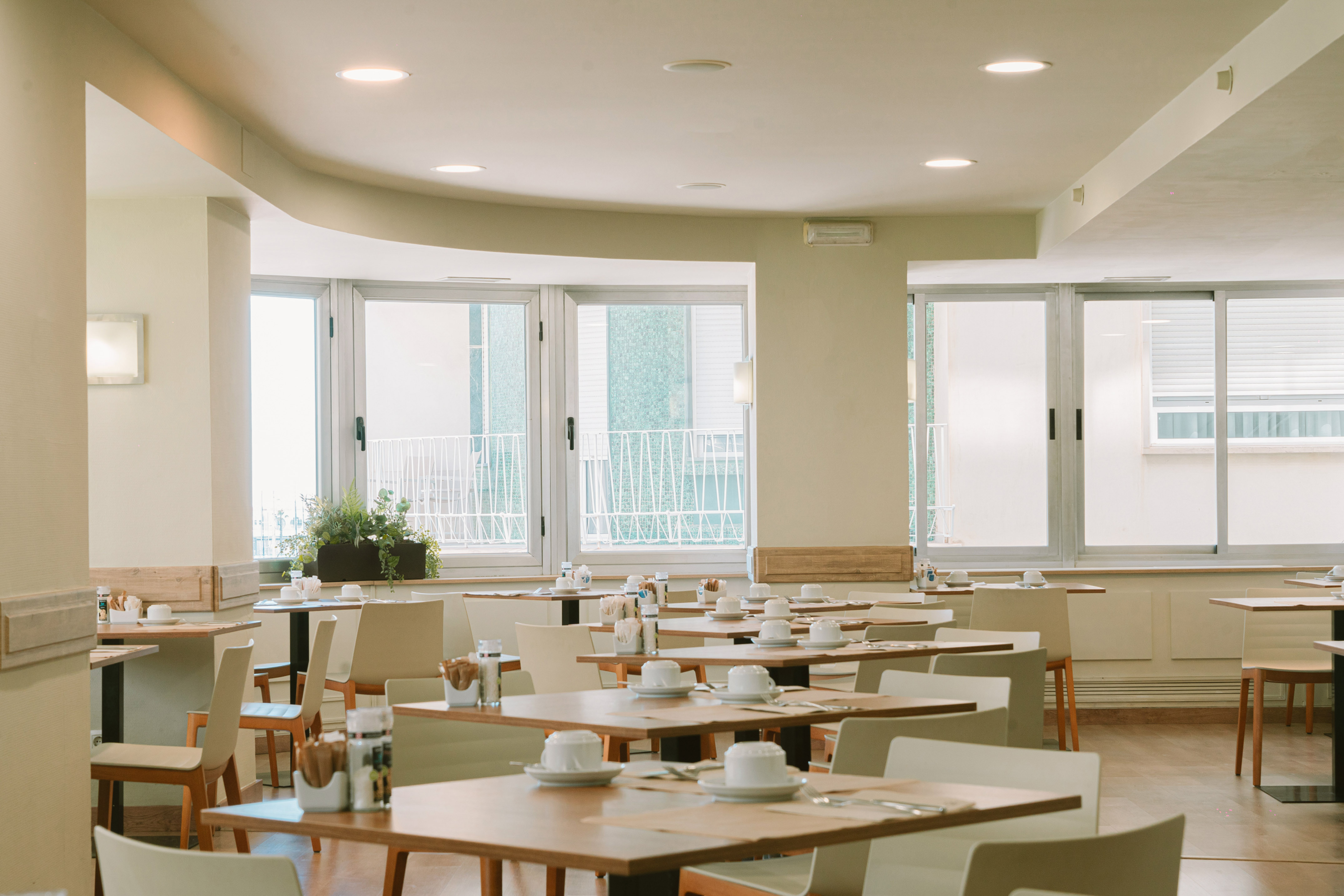Bright hotel dining room with many set tables, chairs, and large windows.